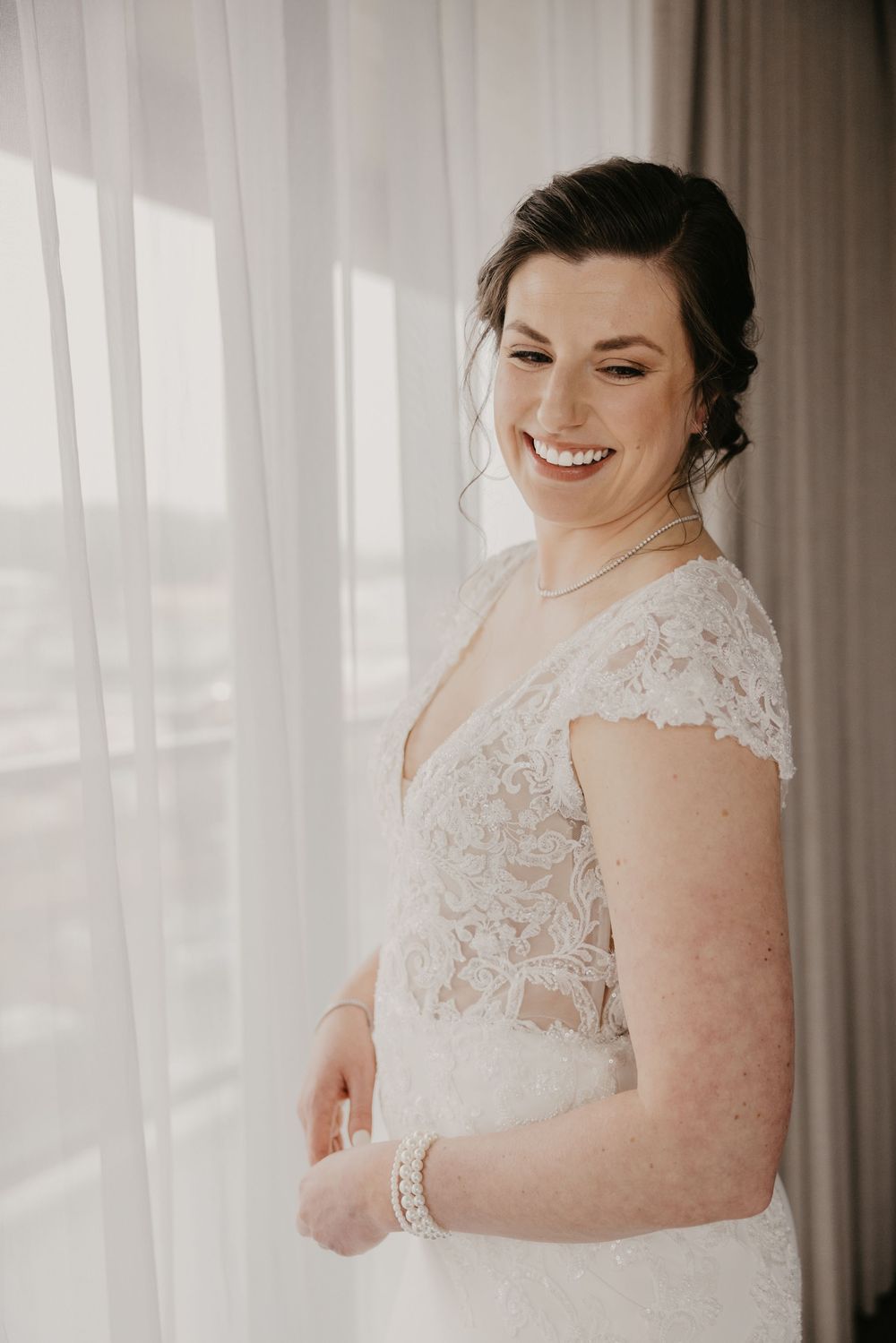 Radiant bride in lace cap-sleeve wedding dress smiling by a sunlit window before ceremony.