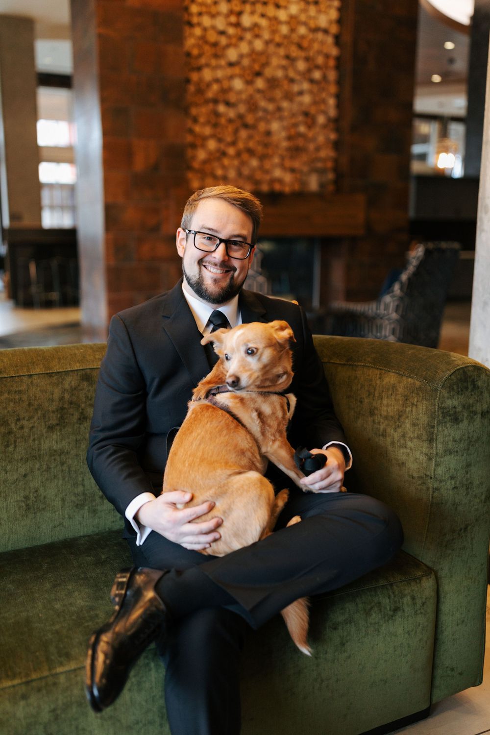 Smiling groom in black suit holding an adorable brown dog on a green velvet sofa.
