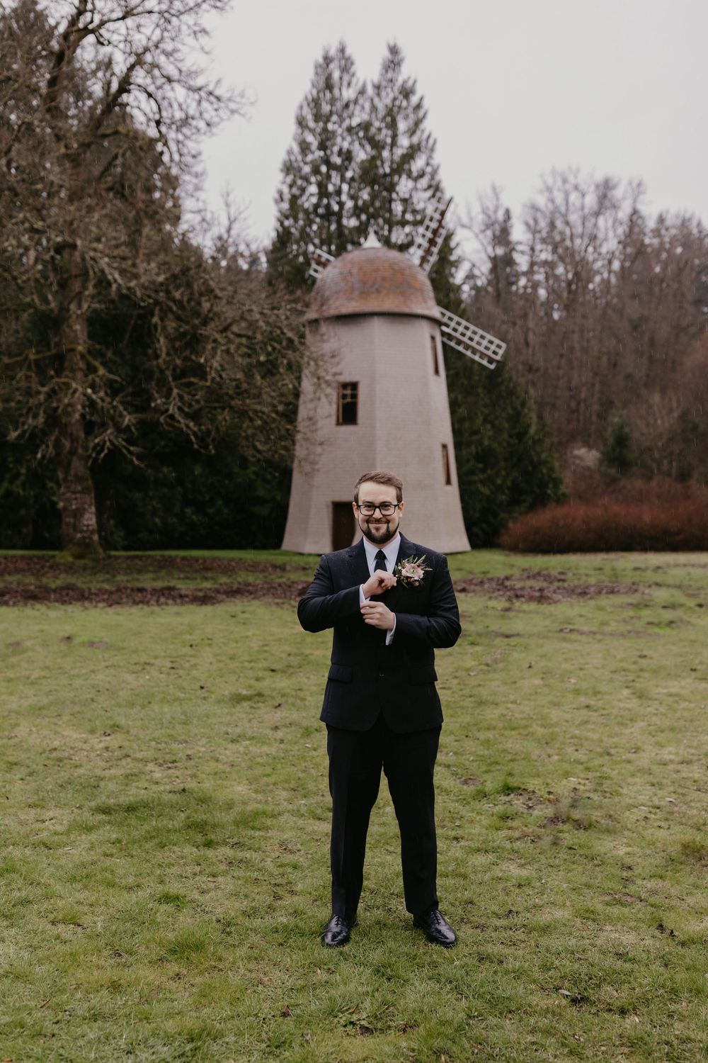 Dapper groom in black suit adjusts cufflinks while standing on green lawn near stone windmill.