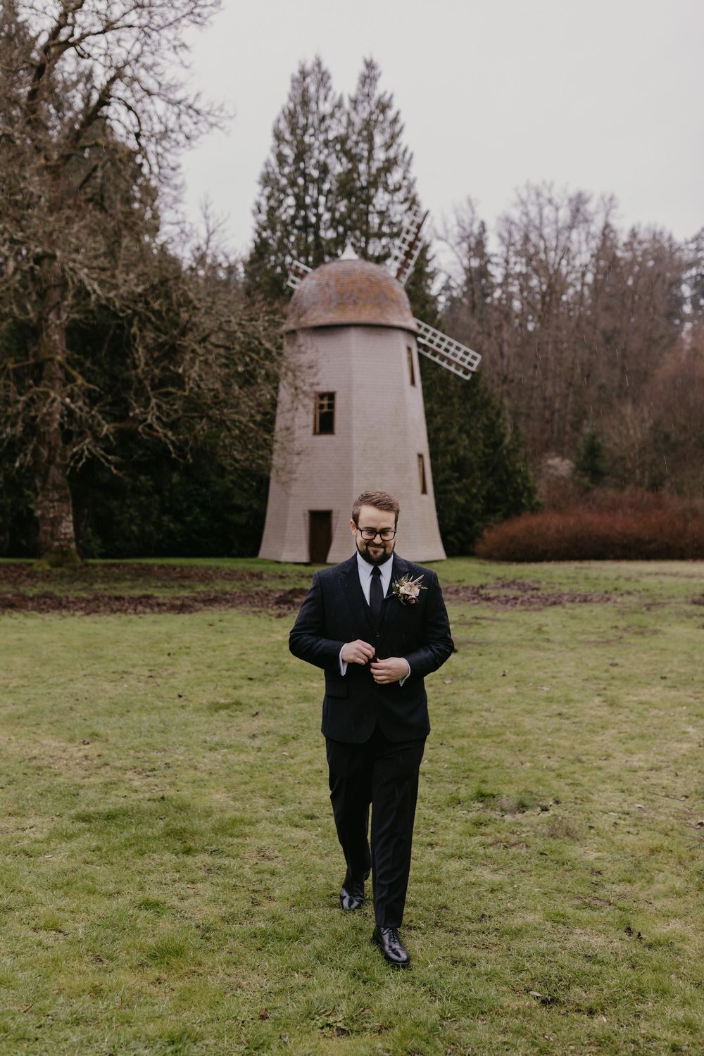Groom in black suit walks across green lawn with a historic windmill in the background.