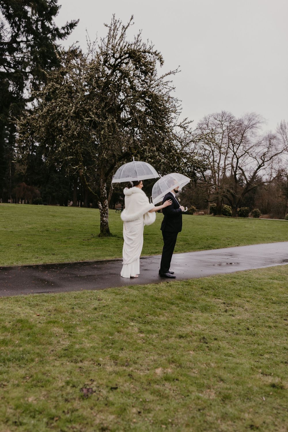 Bride and groom stand facing each other with clear umbrellas on a rainy green garden pathway.