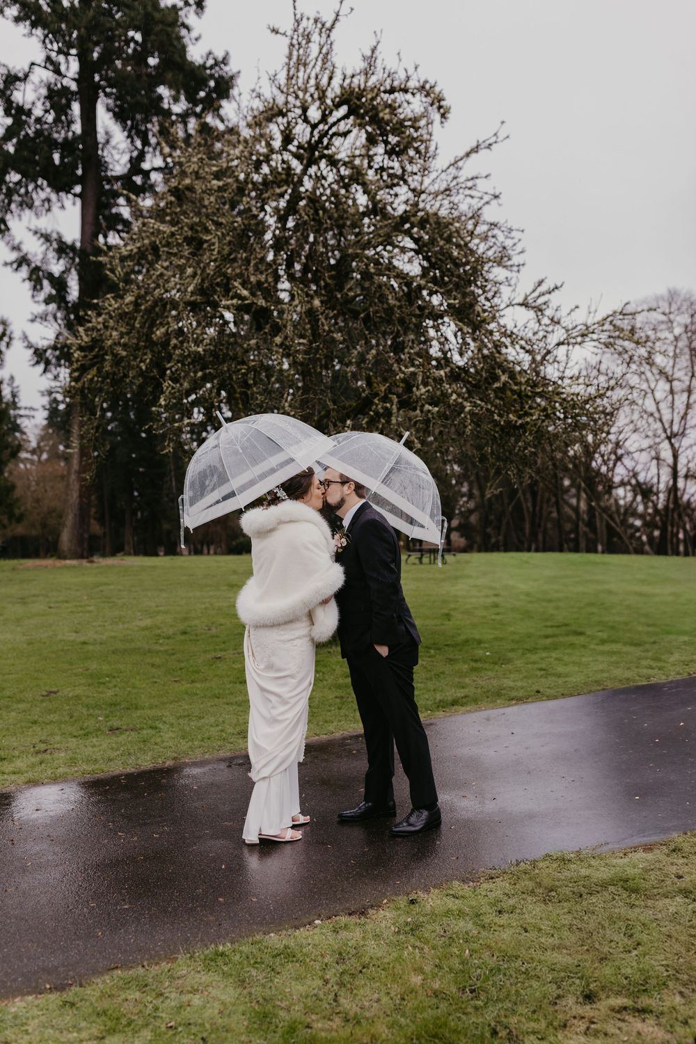 Bride and groom kissing under clear bubble umbrellas on a rainy wedding day in a lush green park.
