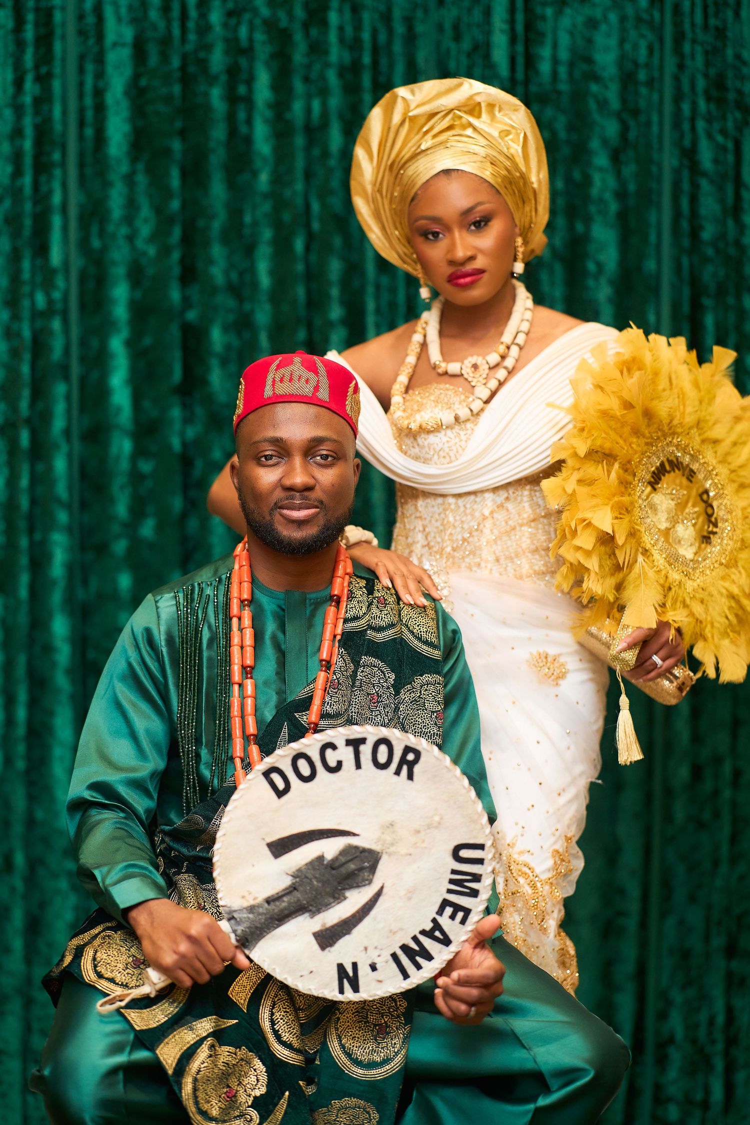 Nigerian couple in traditional Igbo attire, man holding a doctor sign, woman in gold gele headwrap and white lace dress.