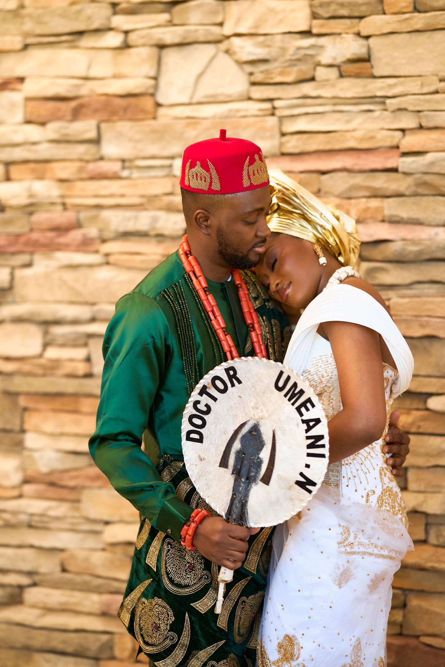 Couple in traditional Nigerian attire embrace, groom holding a fan reading Doctor Umeani N.