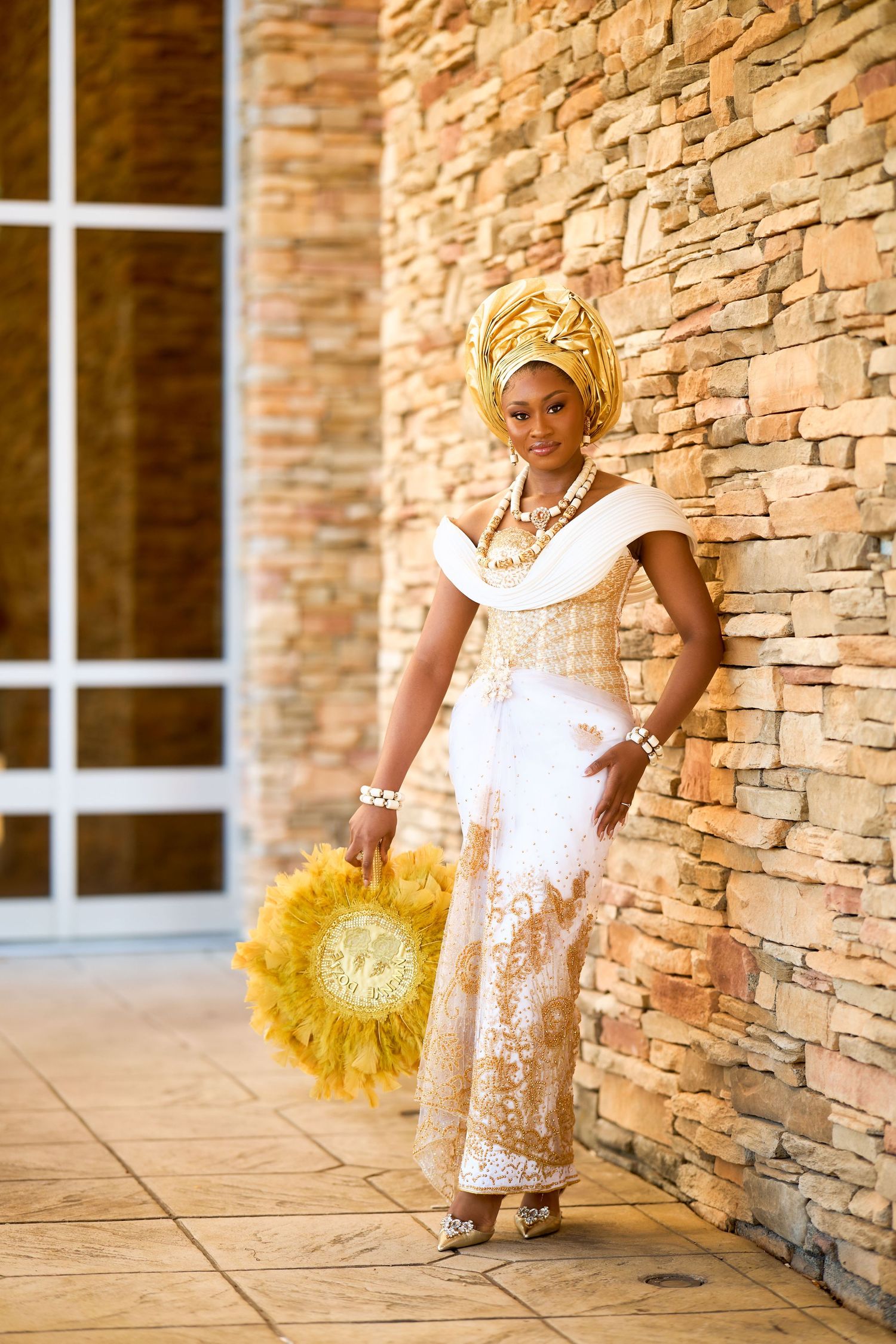 Woman in traditional African attire with gold headwrap, white and gold dress, holding a feathered fan by a stone wall.