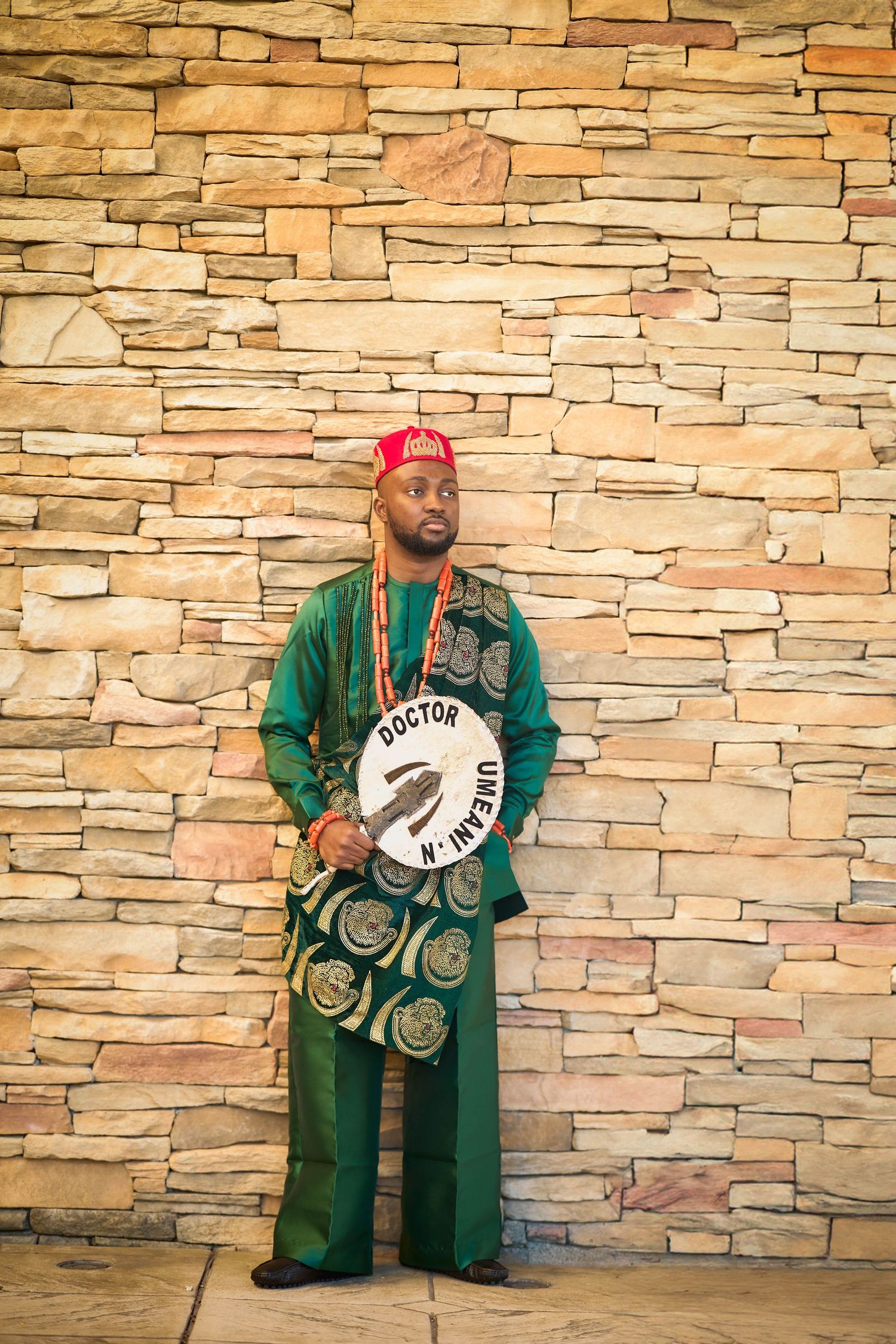 Man in green traditional African attire holding a Doctor Umeali sign against a stone wall.