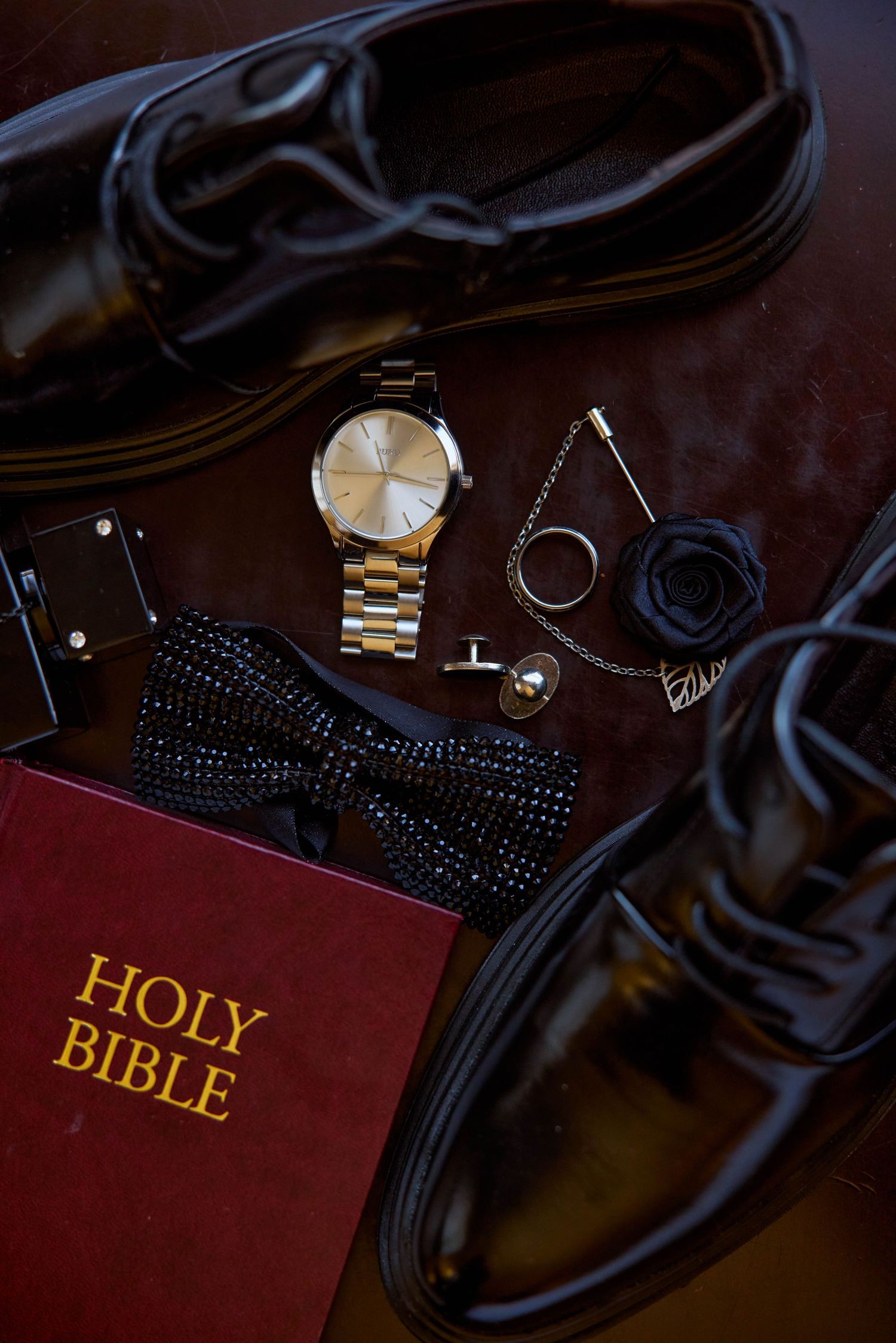 Groom wedding flat lay with Holy Bible, dress shoes, gold watch, bow tie, cufflinks, and accessories on dark surface.