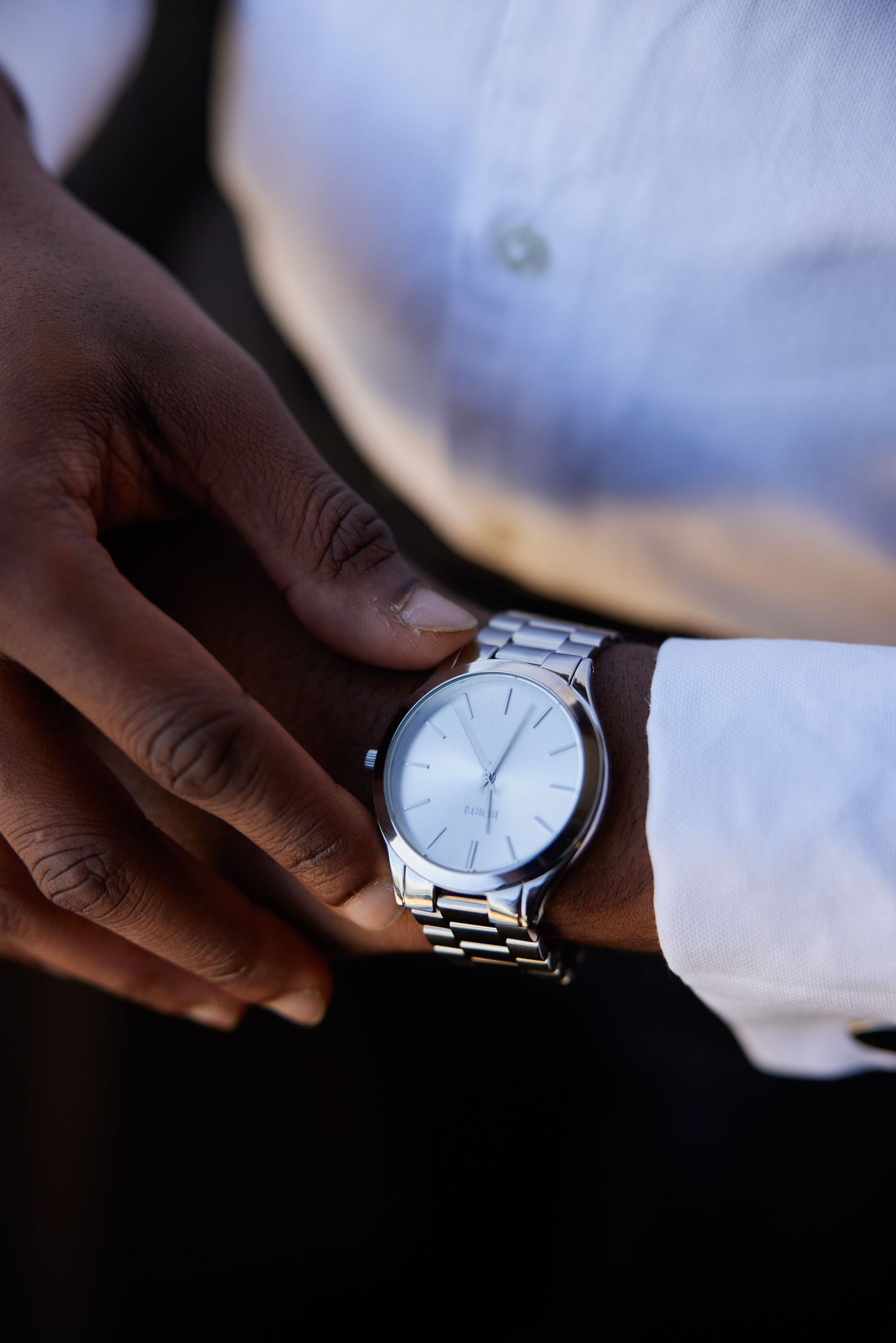 A man wearing a silver analog wrist watch with a minimalist white dial, paired with a white dress shirt.