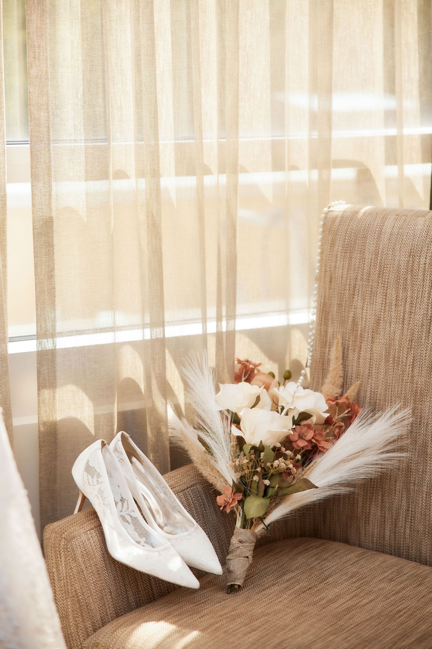 White bridal heels and a boho wedding bouquet with roses and pampas grass rest on a beige armchair by sheer curtains.