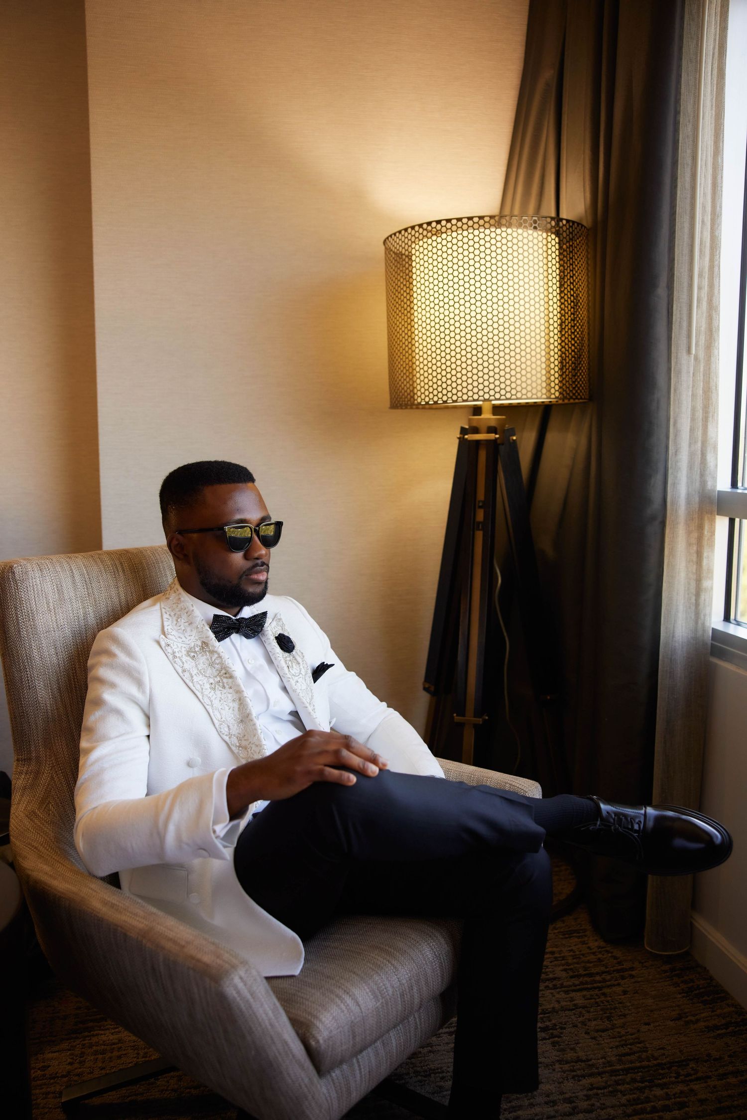 A stylish man in a white tuxedo jacket and bow tie sits in an armchair wearing sunglasses beside a gold lamp.