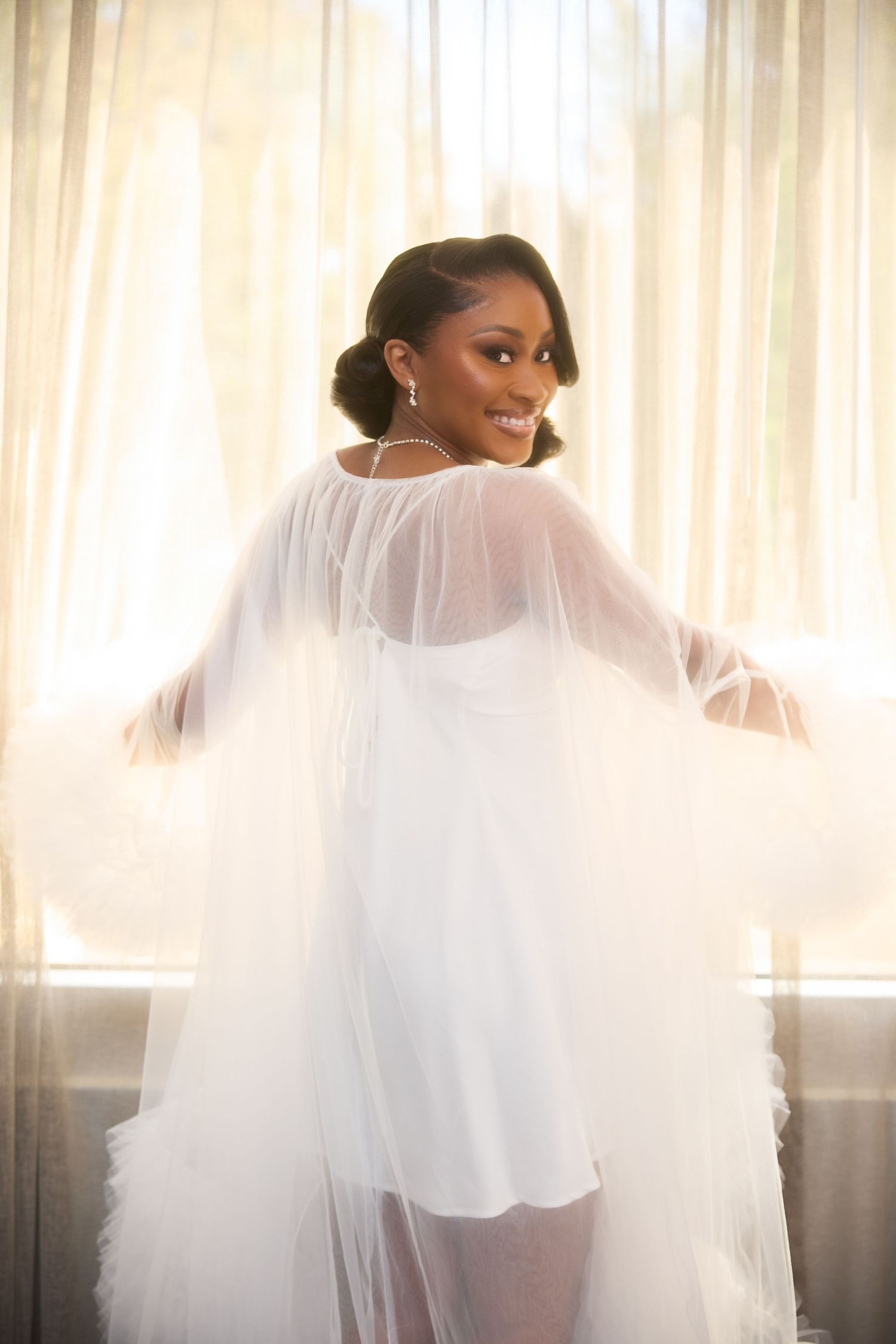Smiling bride in white sheer tulle robe posing elegantly by a sunlit window on her wedding day.