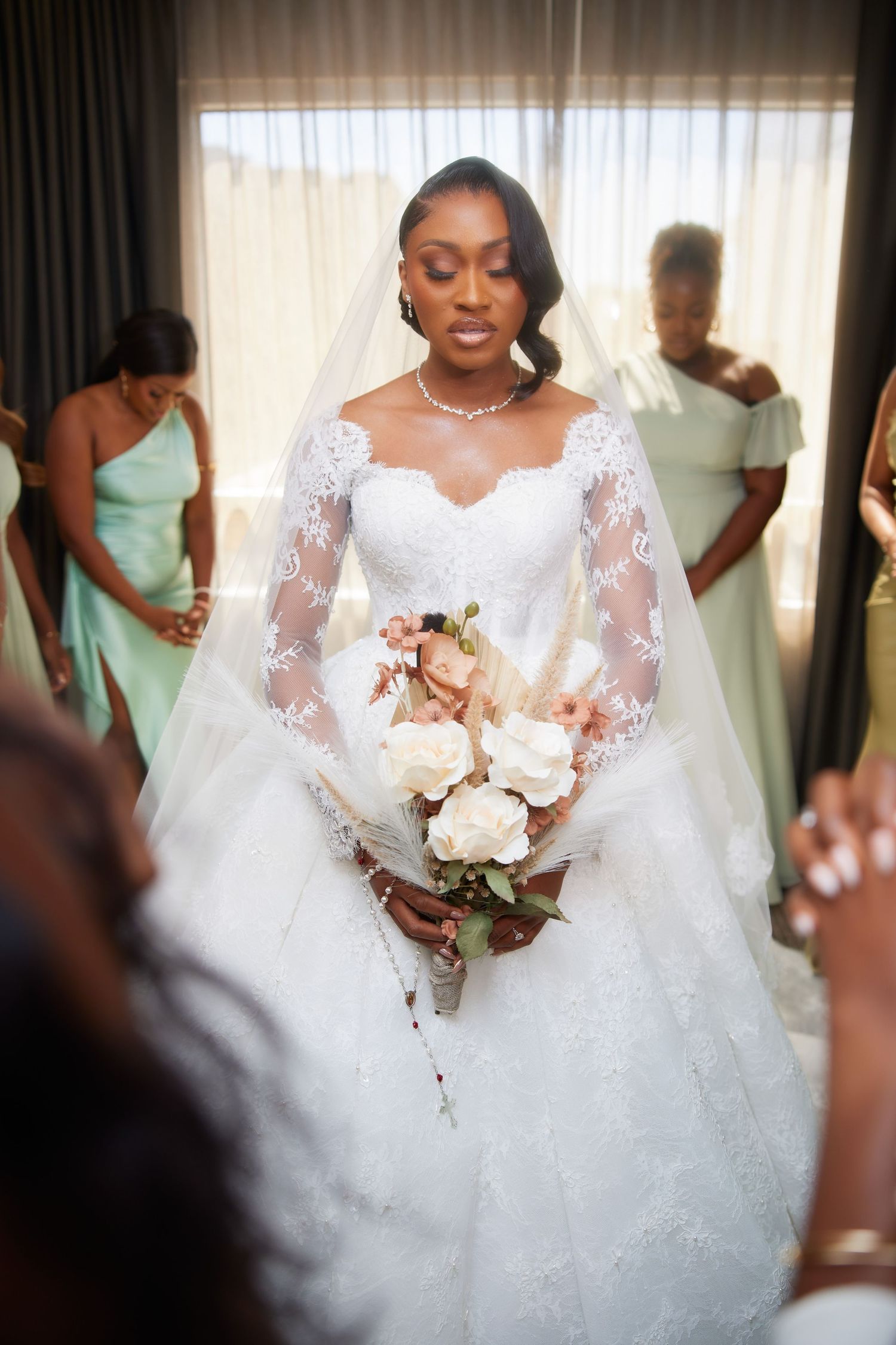 Bride in lace wedding gown holds floral bouquet while bridesmaids in mint dresses bow heads in prayer behind her.