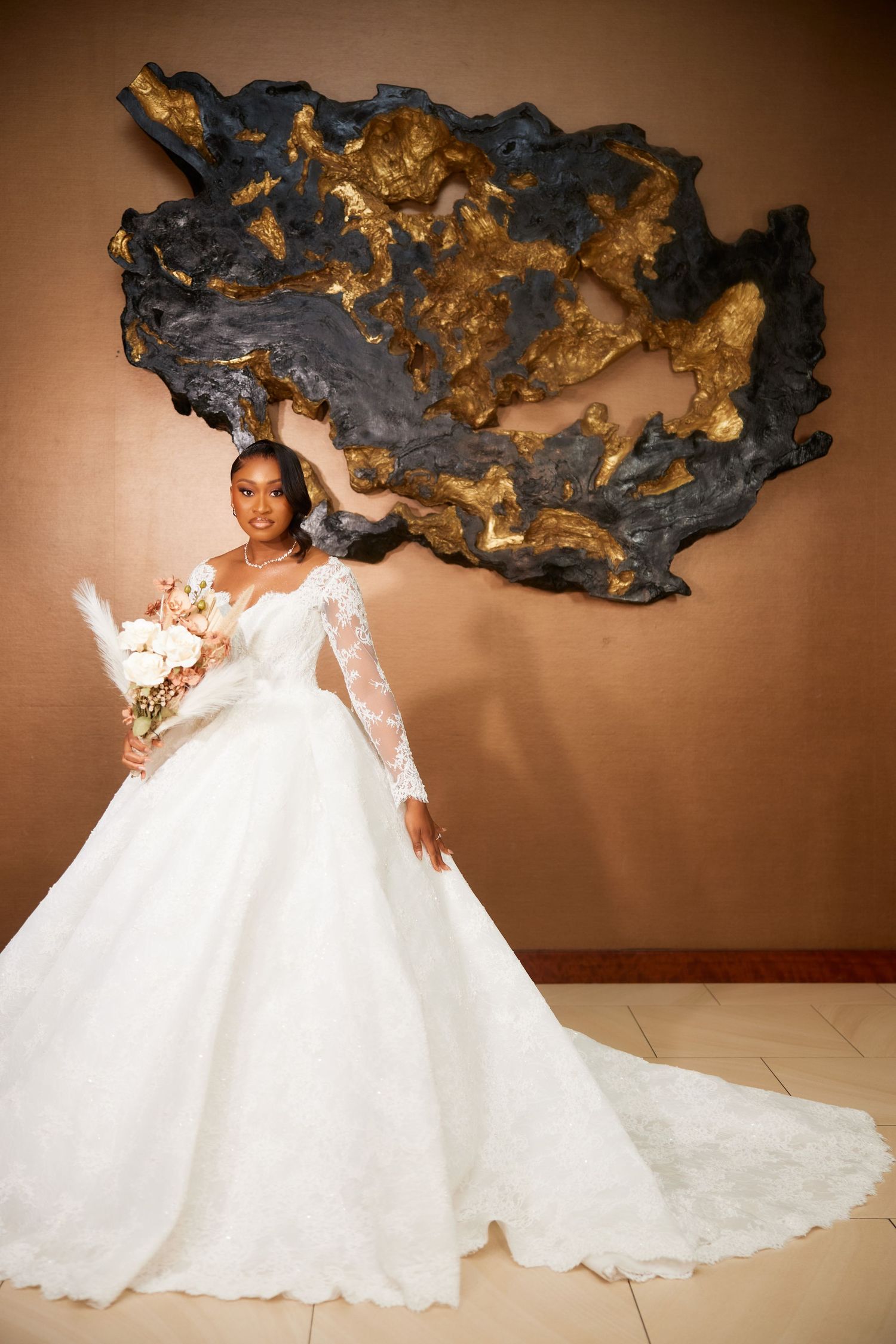Elegant bride in a white lace ball gown with long sleeves holding a bouquet, posing before a gold and black wall art.