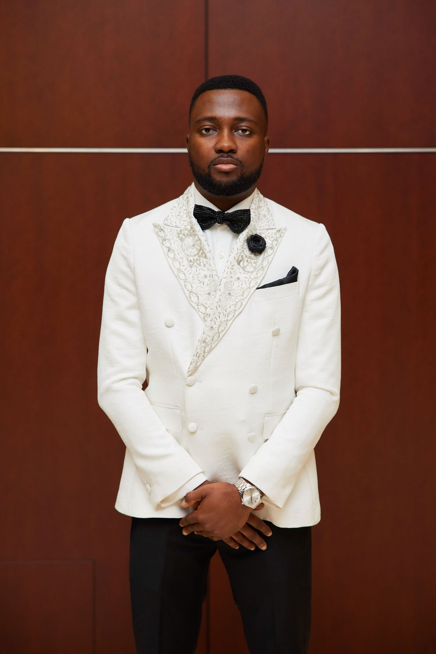 Man in white embroidered tuxedo jacket with black bow tie and pocket square posing formally.