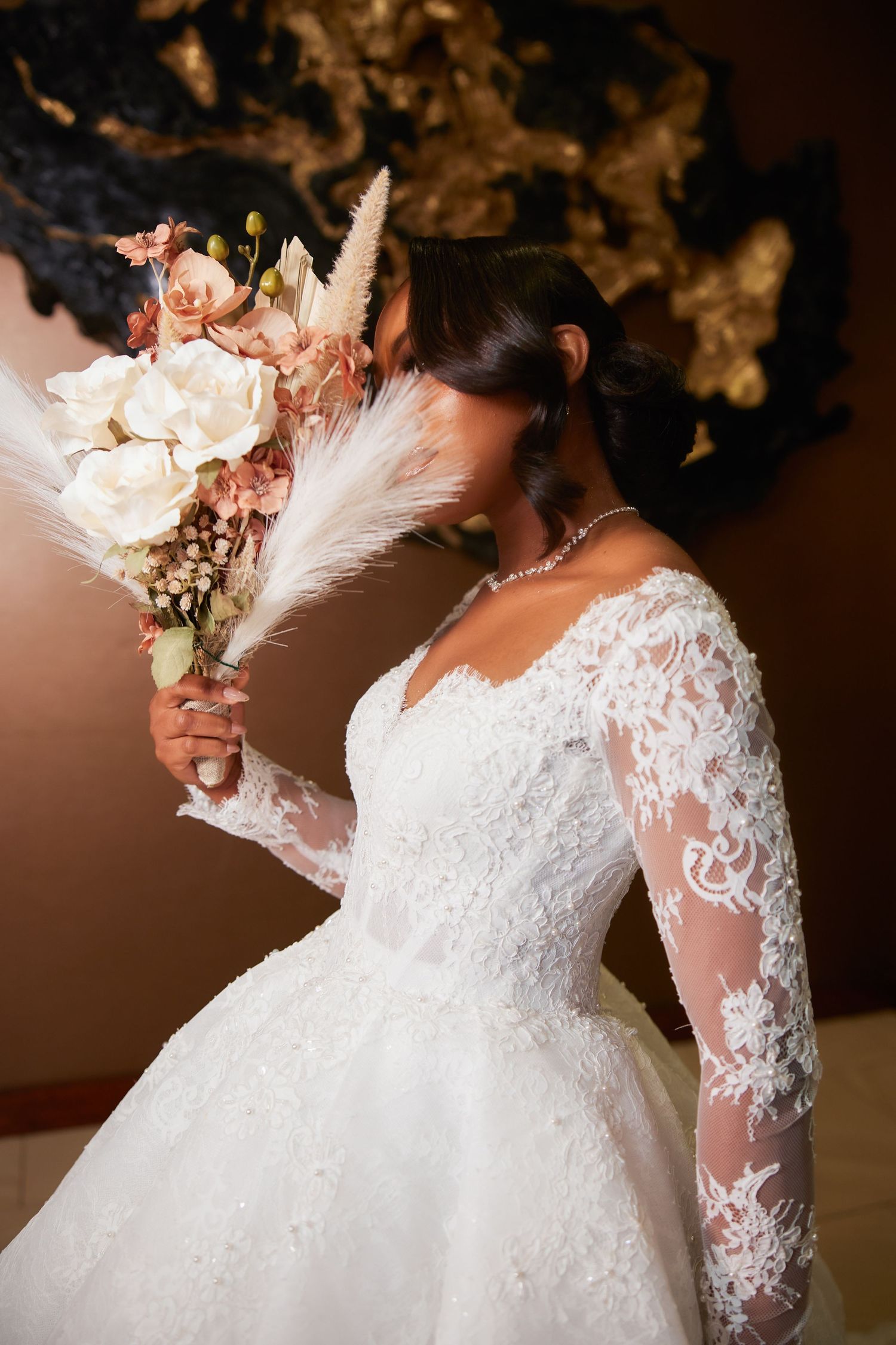 Bride in lace long-sleeve wedding gown holding a boho bouquet with white roses and pampas grass, face hidden by flowers.