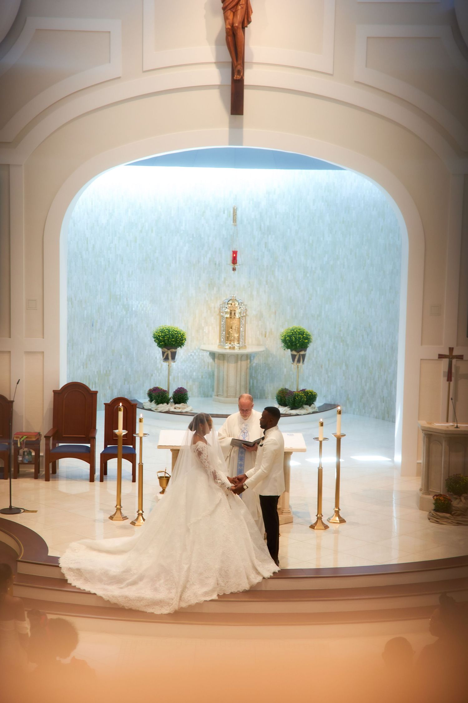 Bride and groom exchanging vows at a church altar with a priest, surrounded by candles and floral arrangements.
