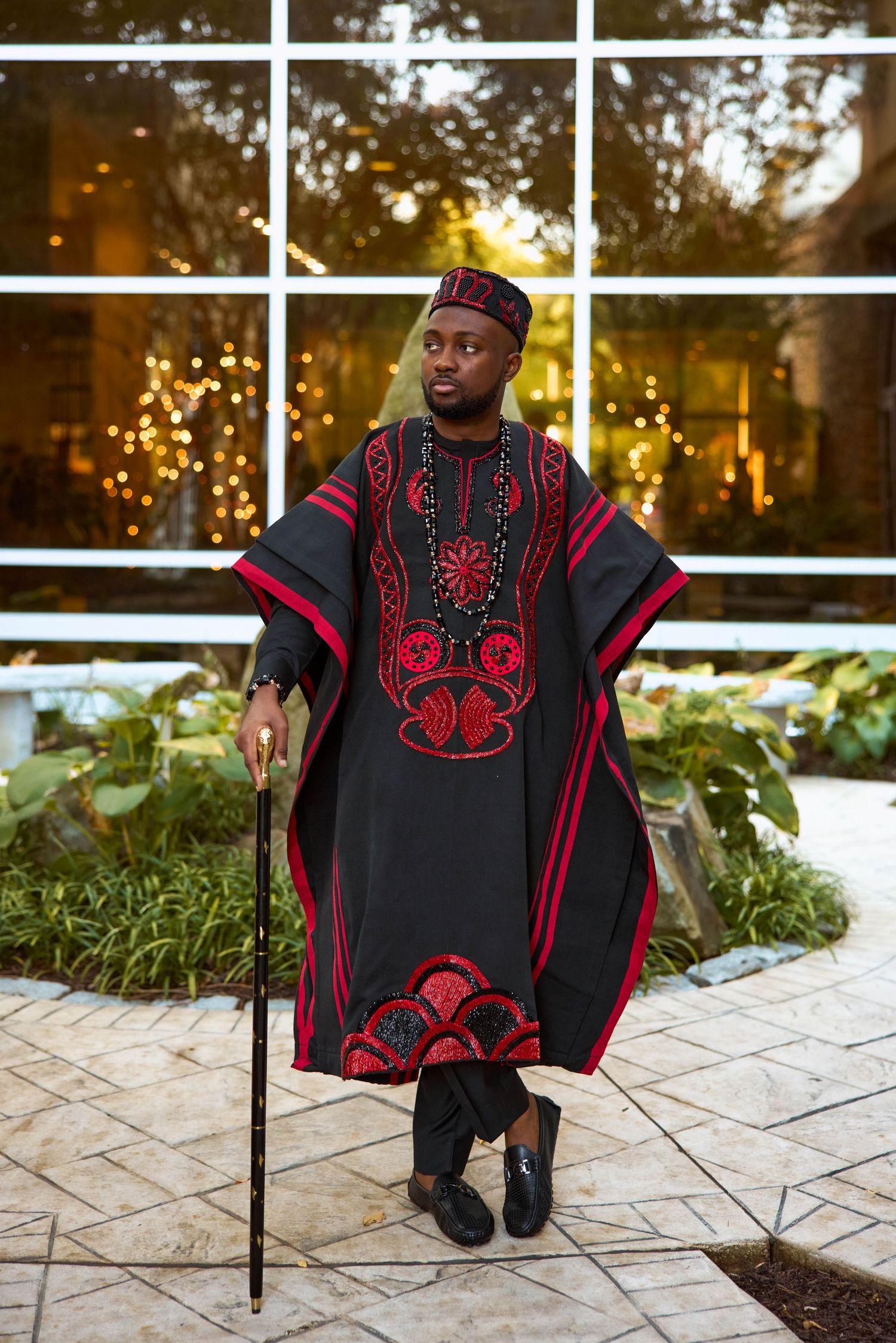 Man in black and red African dashiki with matching cap holds a cane, posing outdoors near glass building.
