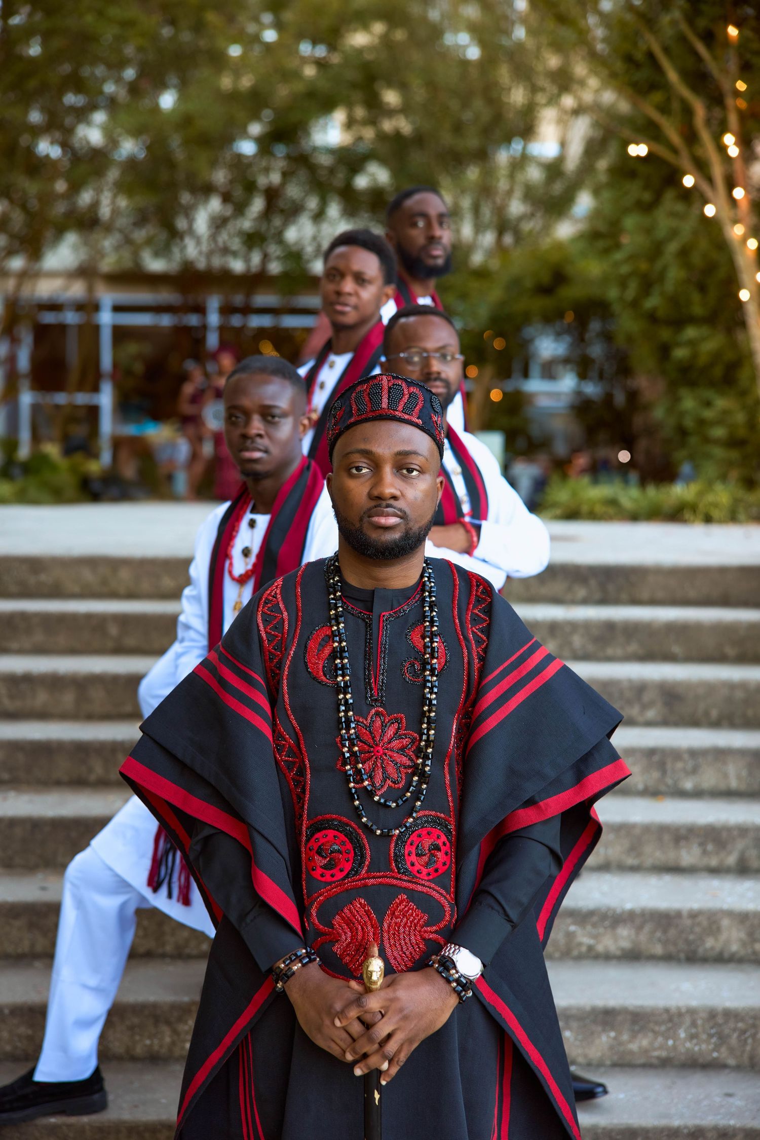 Men in traditional African attire with black and red embroidered robes pose on outdoor steps for a formal occasion.