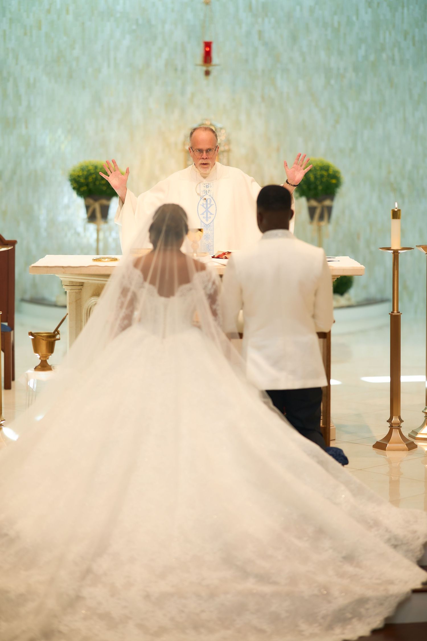 Bride and groom kneeling at church altar before priest with raised hands during Catholic wedding ceremony.