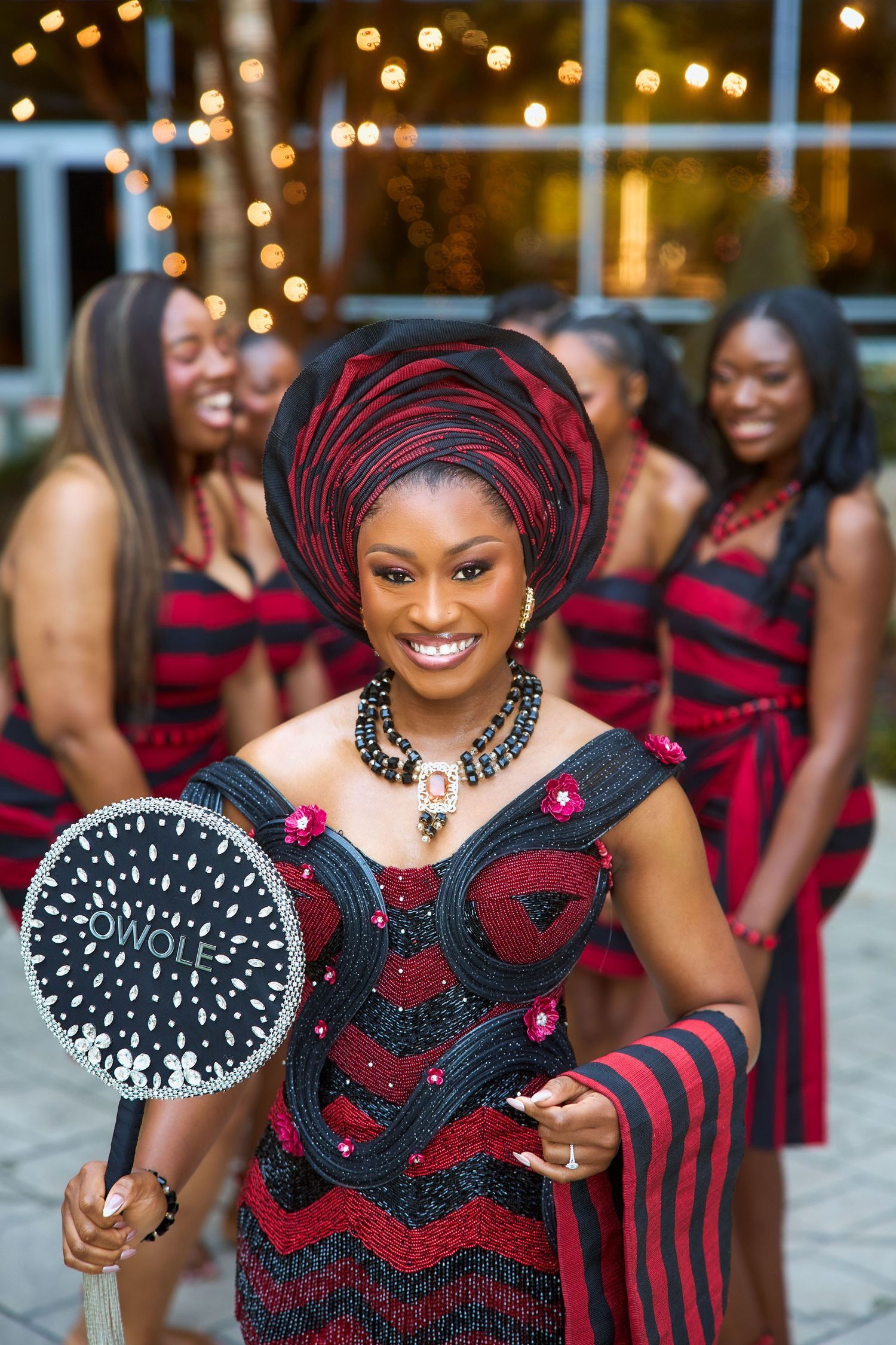 Woman in red and black African traditional attire holding a decorative fan, with bridesmaids in matching outfits behind her.
