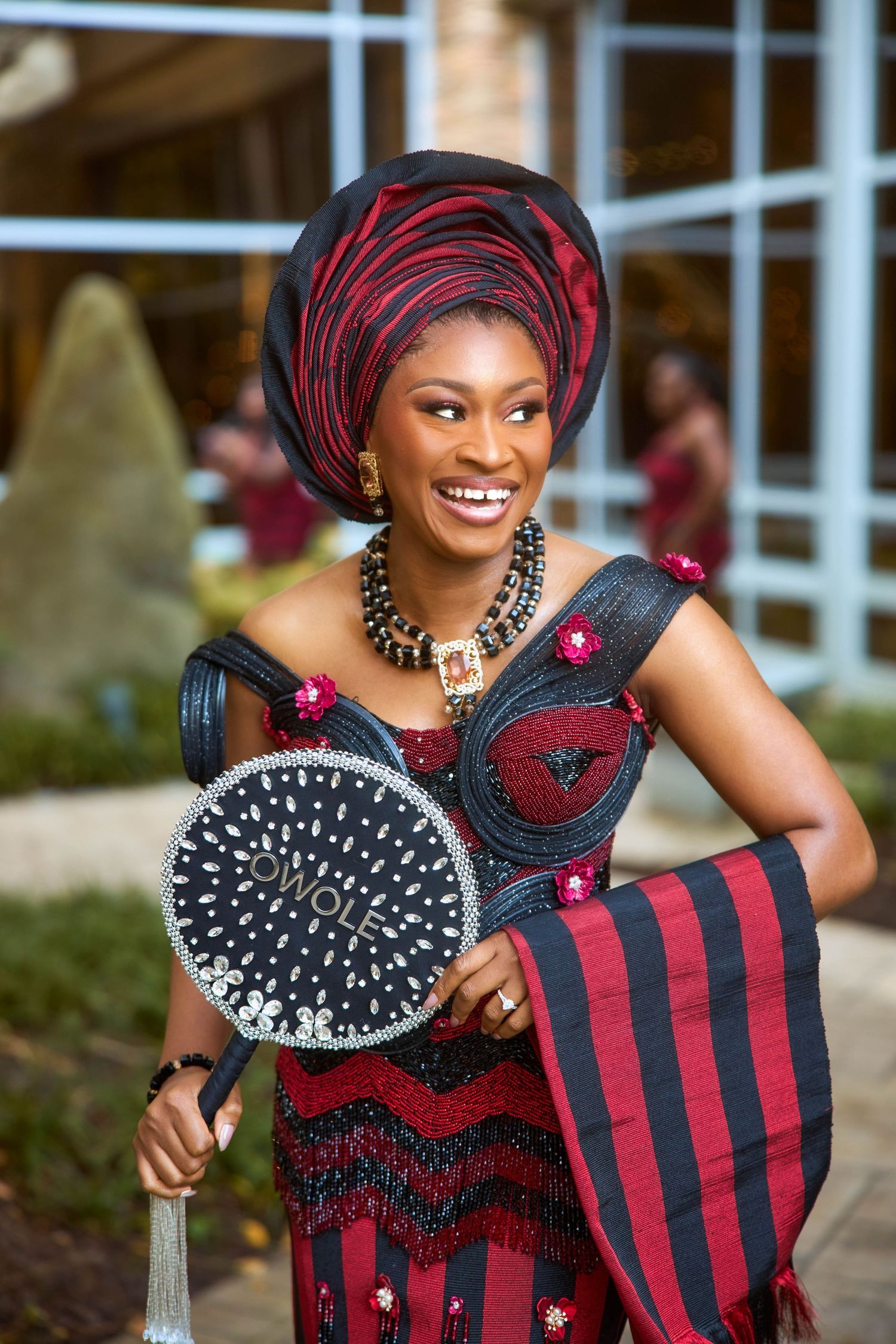 Smiling woman in vibrant red and black African traditional attire, gele headwrap, beaded jewelry, holding decorative fan.