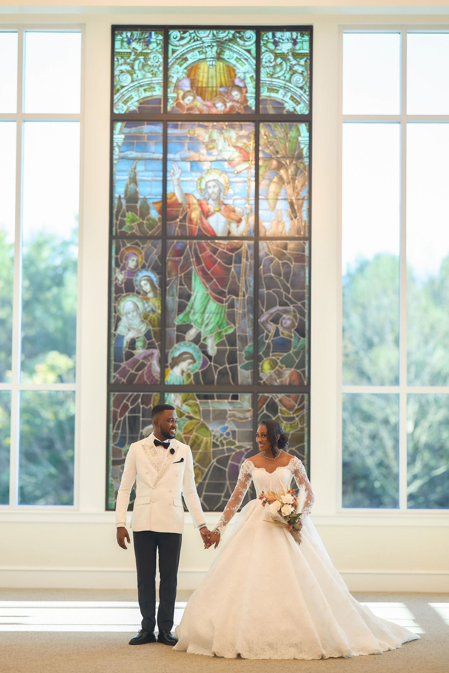 Bride and groom hold hands before a stunning stained glass window in a sunlit chapel on their wedding day.