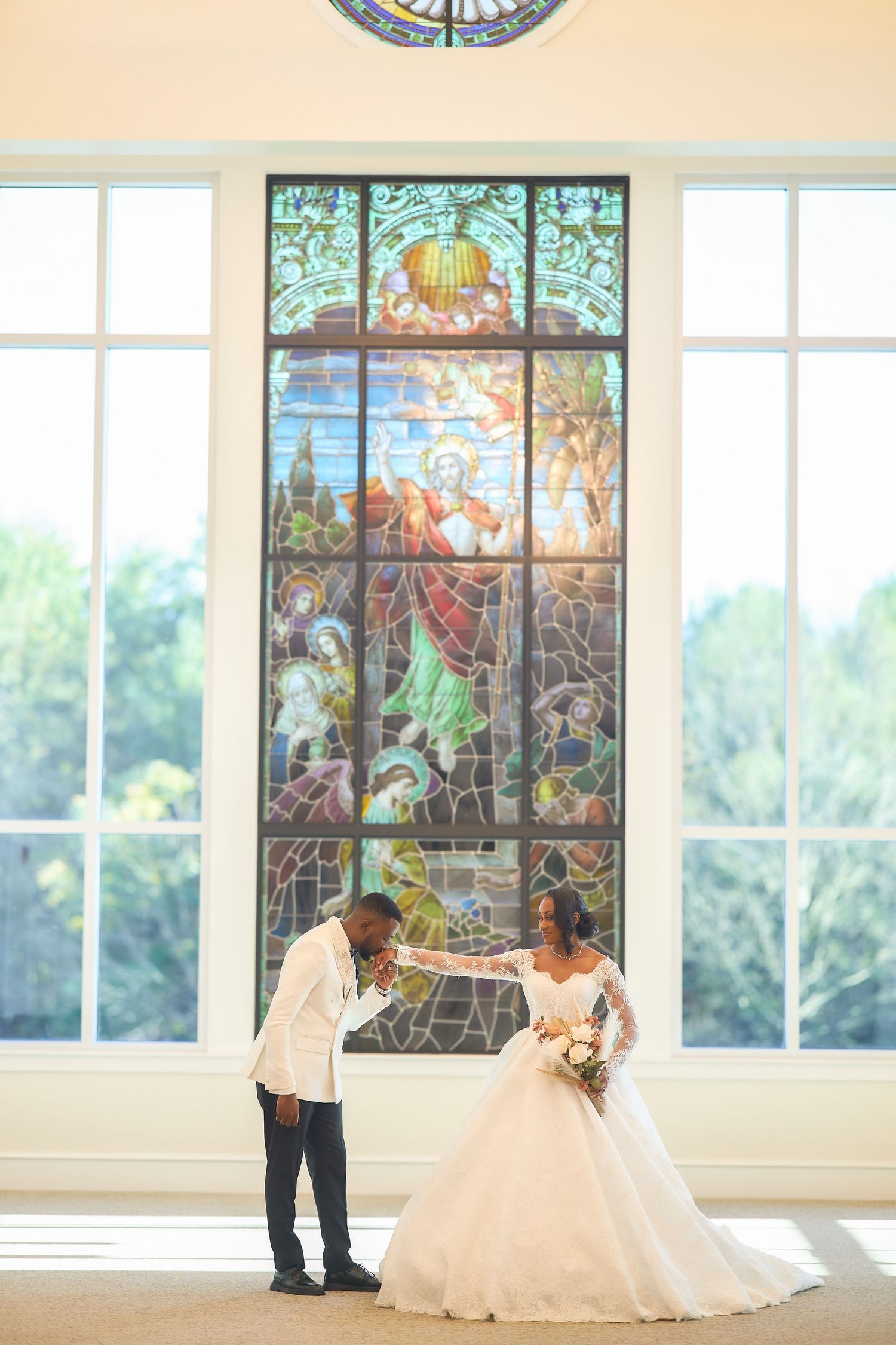 Bride and groom pose before a stunning stained glass window in a bright chapel, bride in a ball gown holding a bouquet.