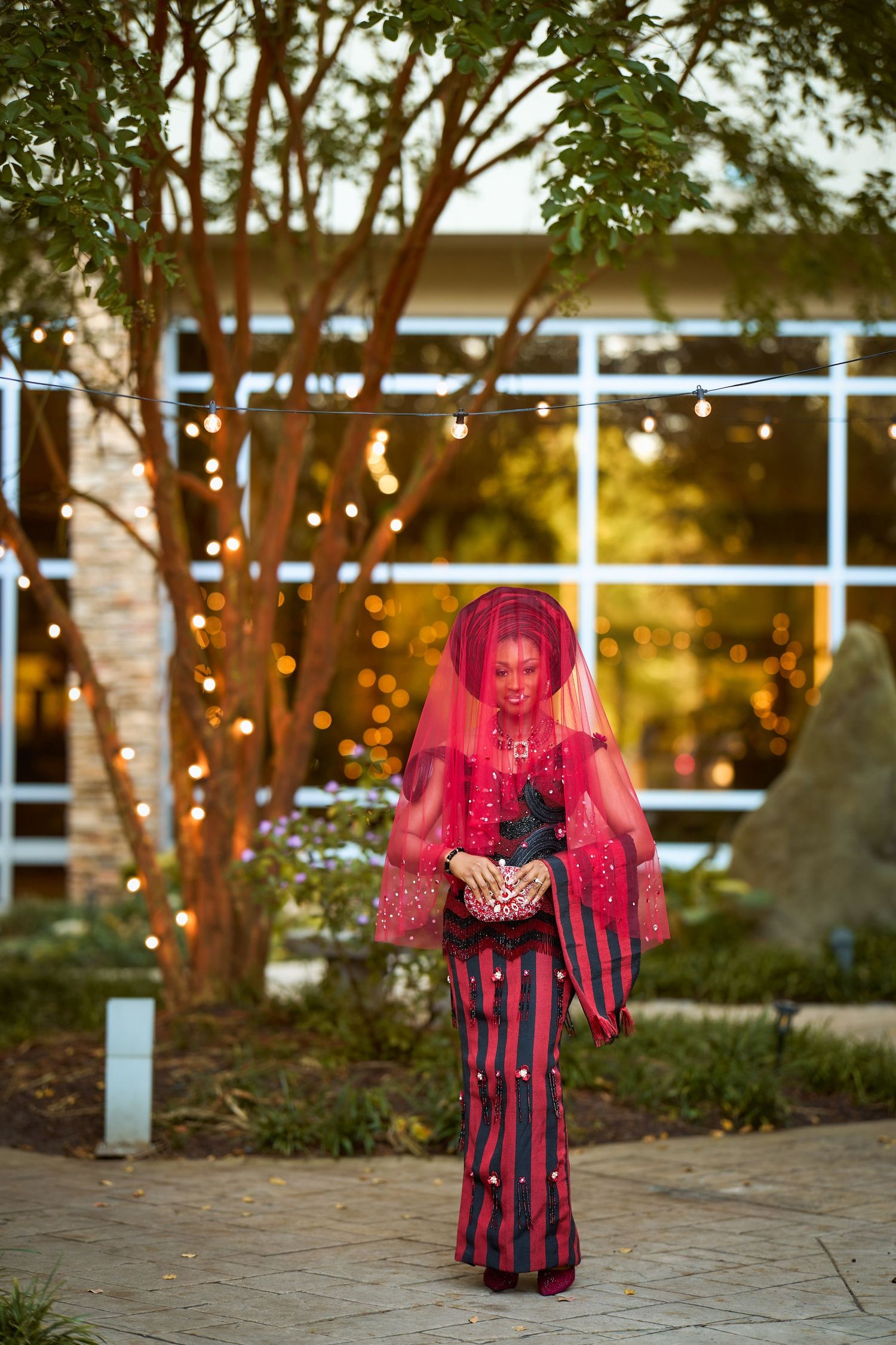 Woman in red veil and striped outfit stands outdoors near trees with string lights at dusk.