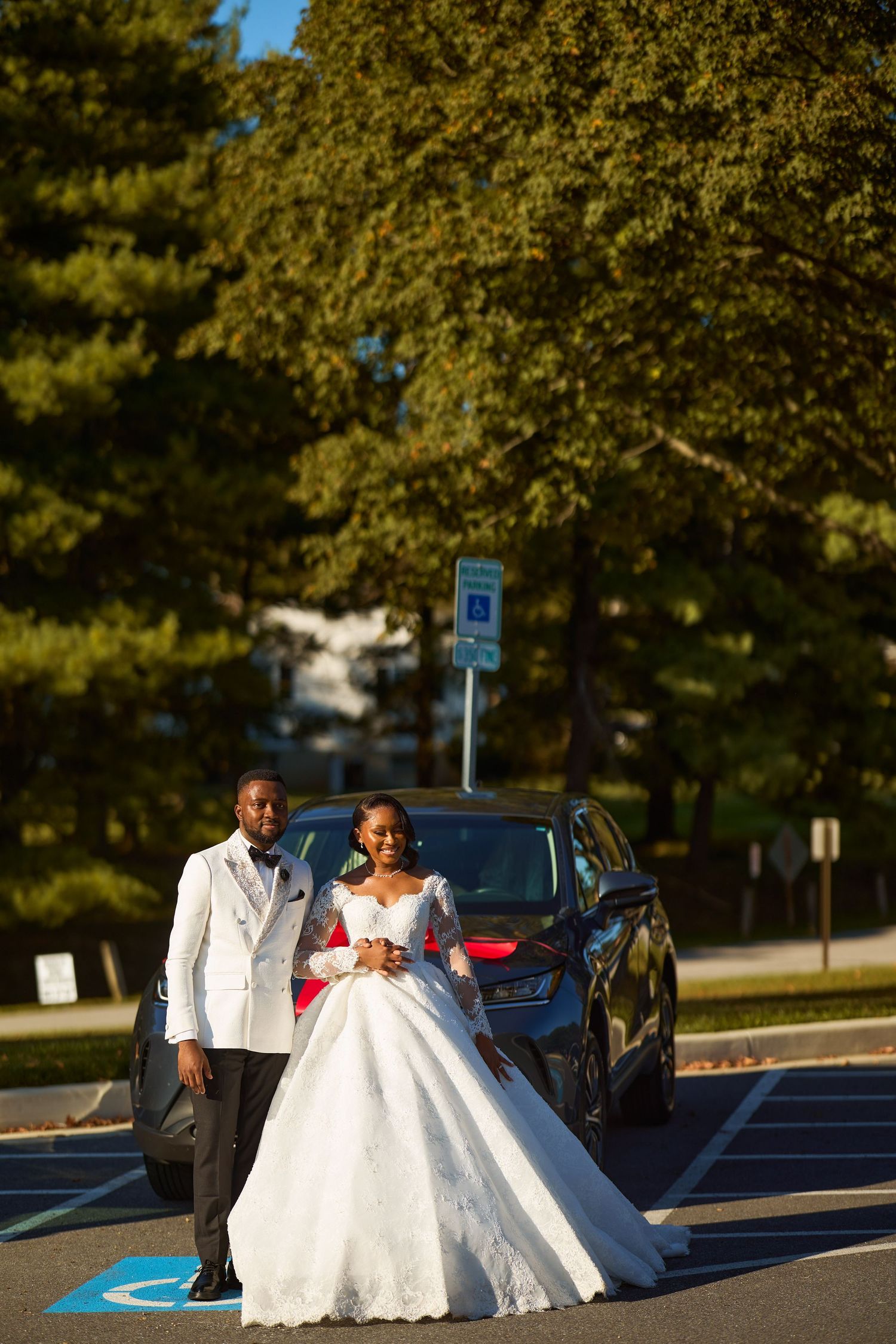 Elegant bride and groom in wedding attire posing in front of a dark car in a sunny parking lot.