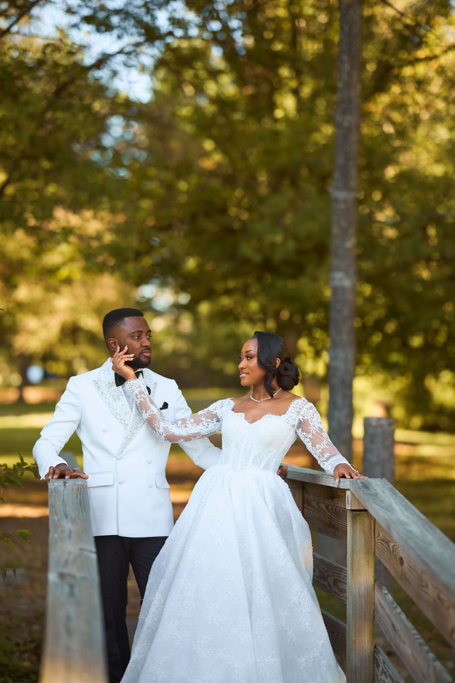 Bride in lace gown and groom in white tuxedo pose on wooden fence in lush green park setting.