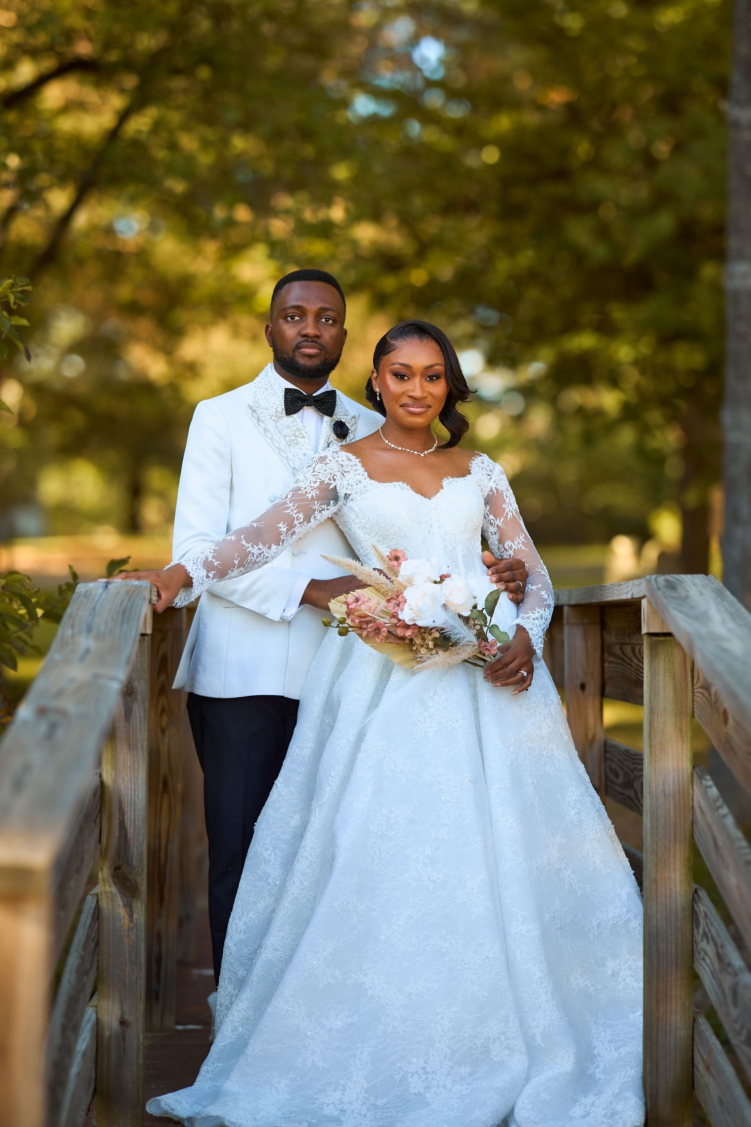 A Black couple poses on a wooden bridge in wedding attire, bride holding a bouquet, surrounded by autumn trees.
