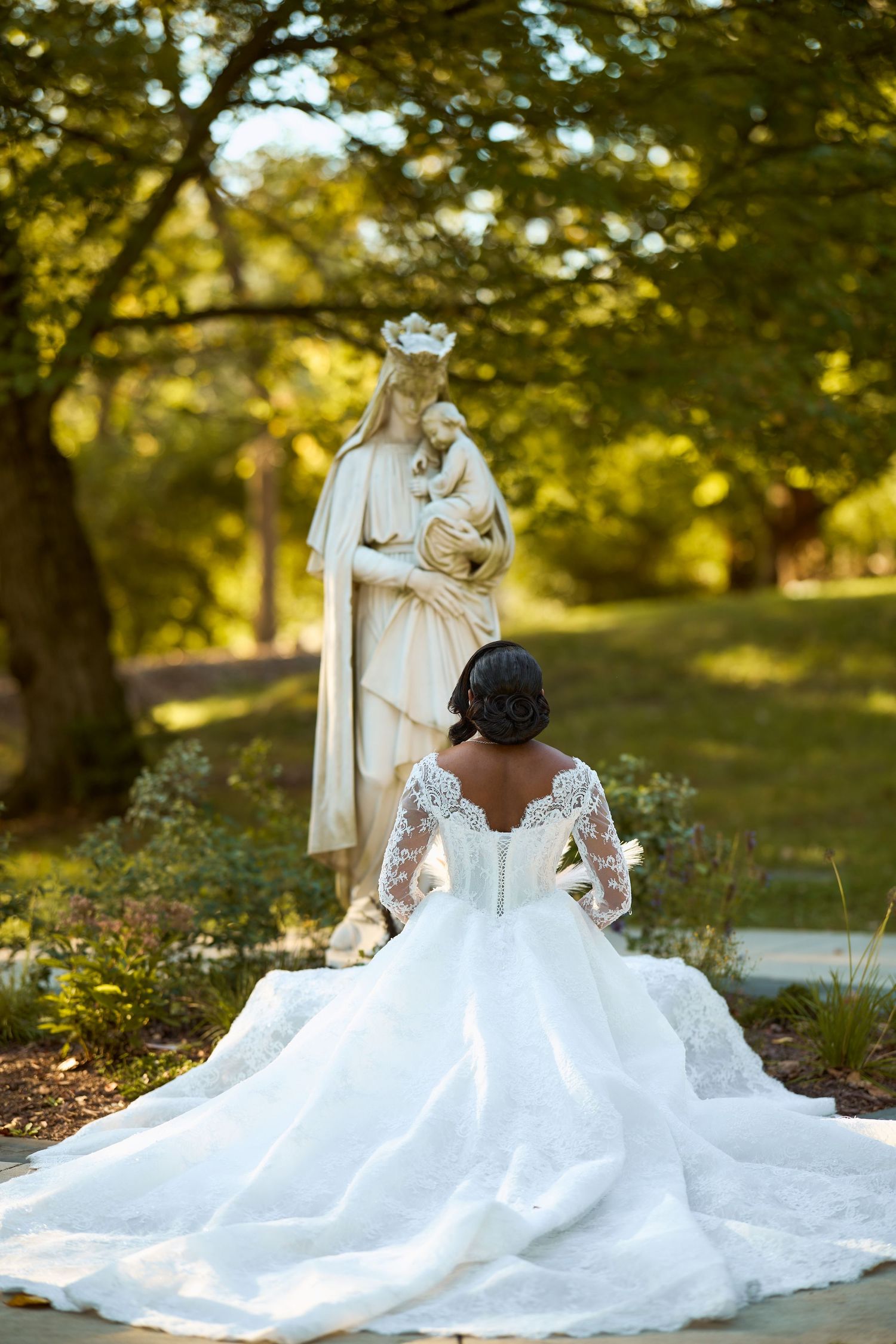Bride in lace wedding gown kneeling before Virgin Mary statue in lush garden setting.