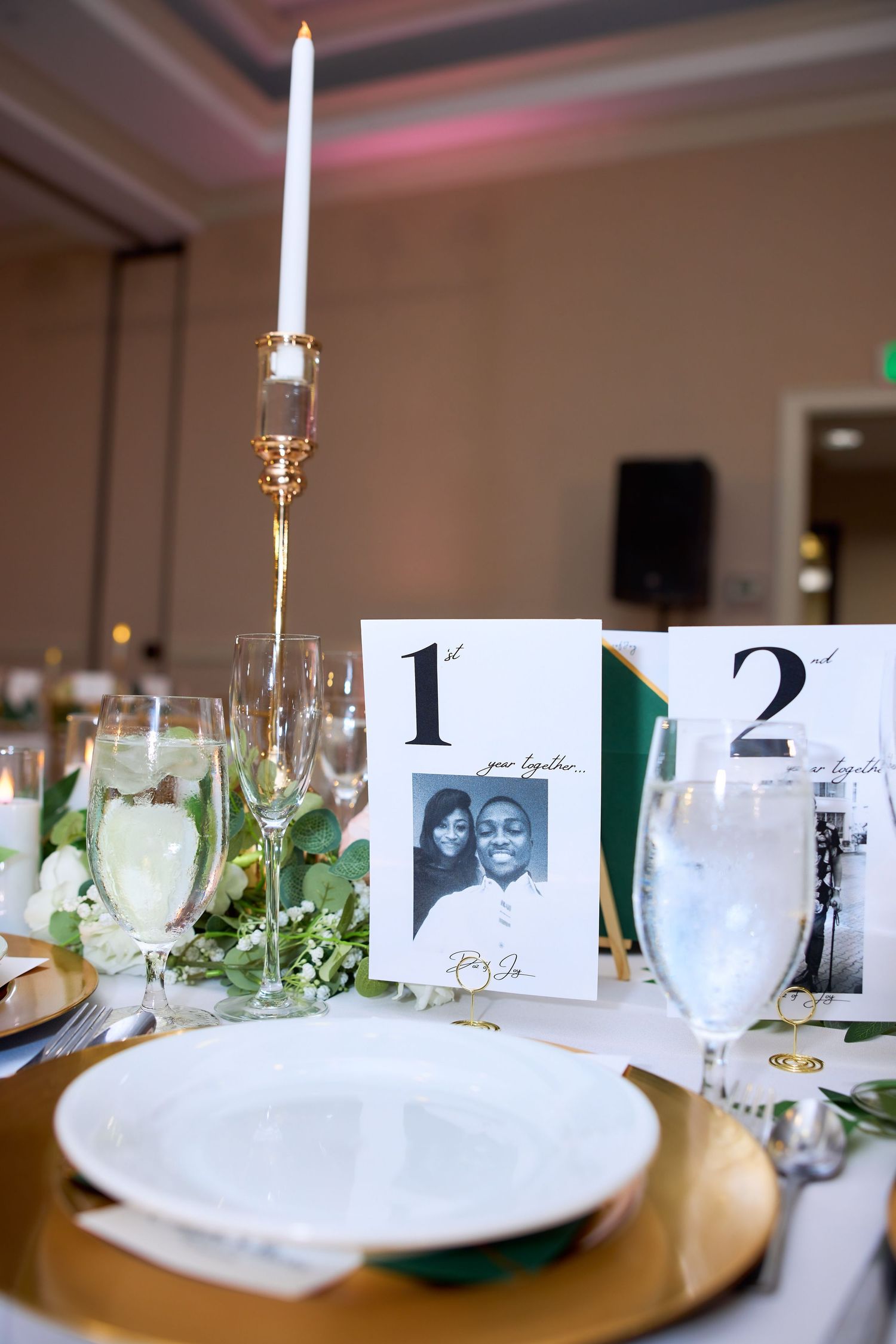 Elegant wedding reception table with gold chargers, candles, greenery, and personalized photo table numbers on white linen.