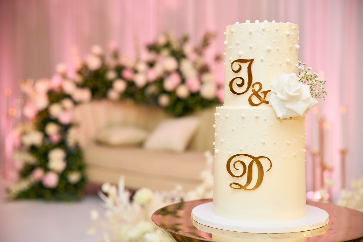 Elegant two-tier white wedding cake with gold monogram initials J and D, adorned with pearl dots and white floral decoration.
