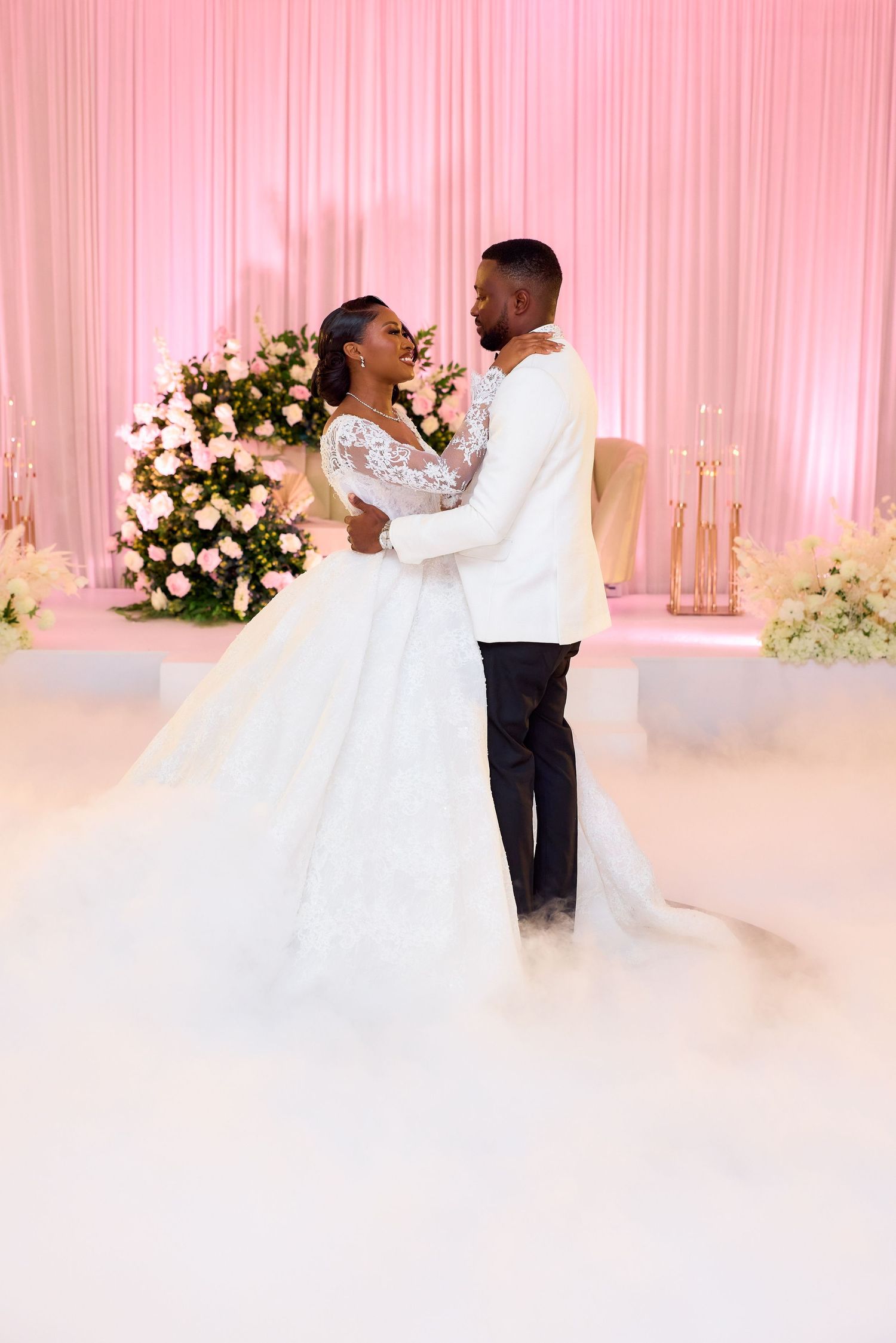 A bride and groom share their first dance surrounded by floral arrangements and soft pink lighting at their elegant wedding reception.