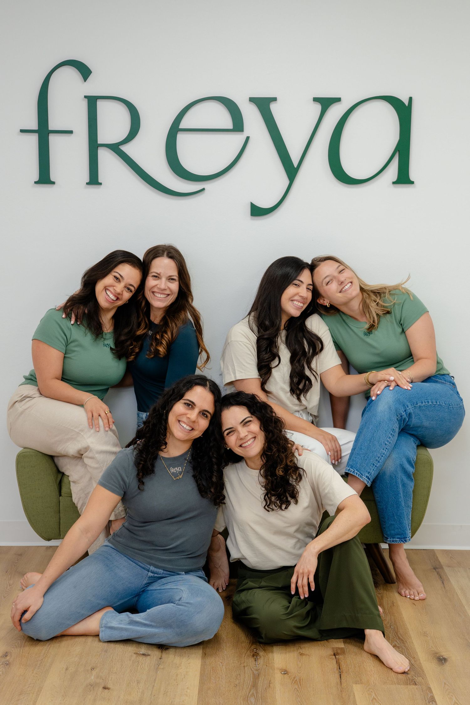 Six women pose together smiling in front of a green Freya logo sign, wearing casual earth-toned outfits.