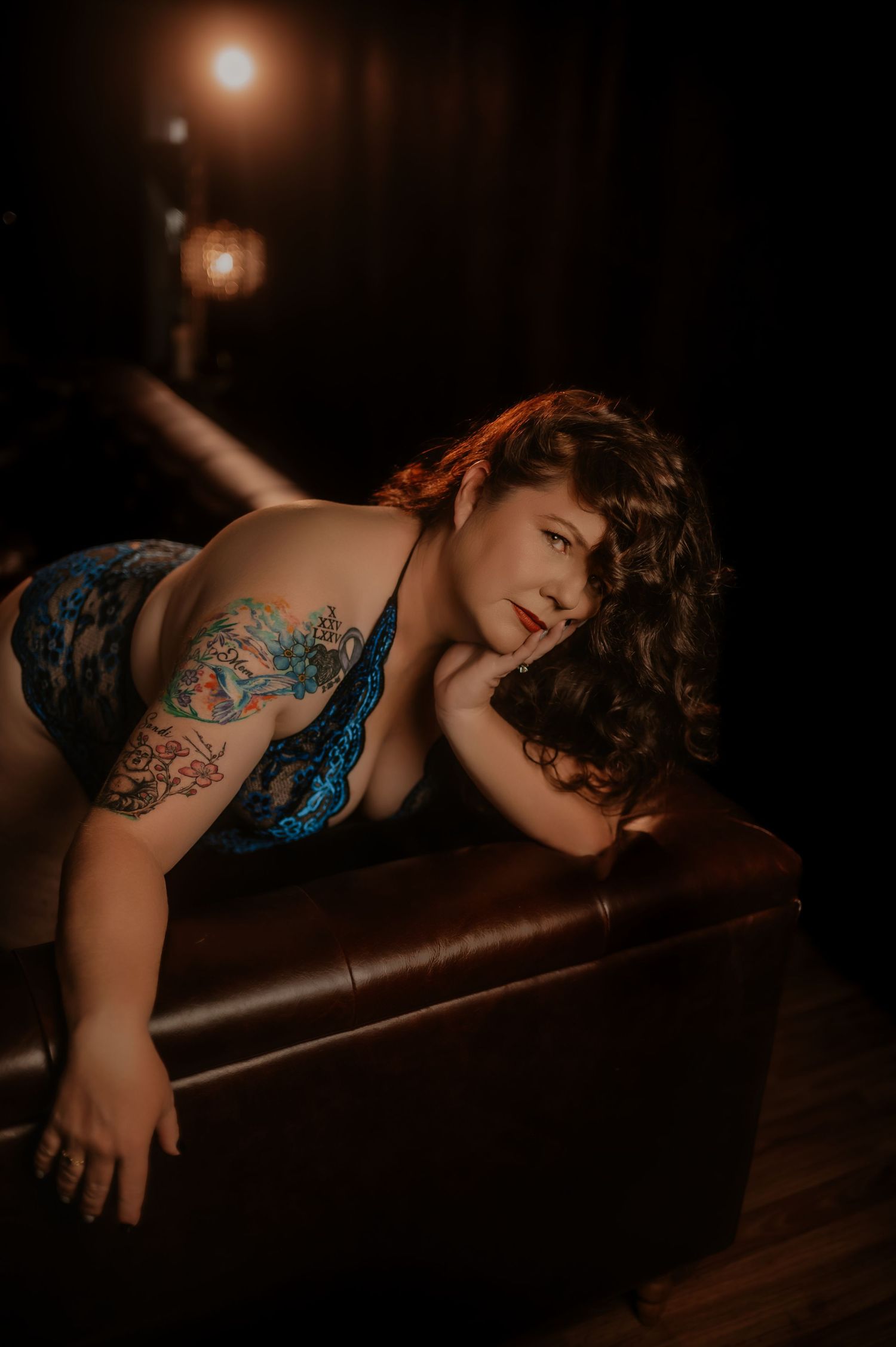 Woman with tattoos in green lingerie lying on dark satin sheets in moody boudoir photography.