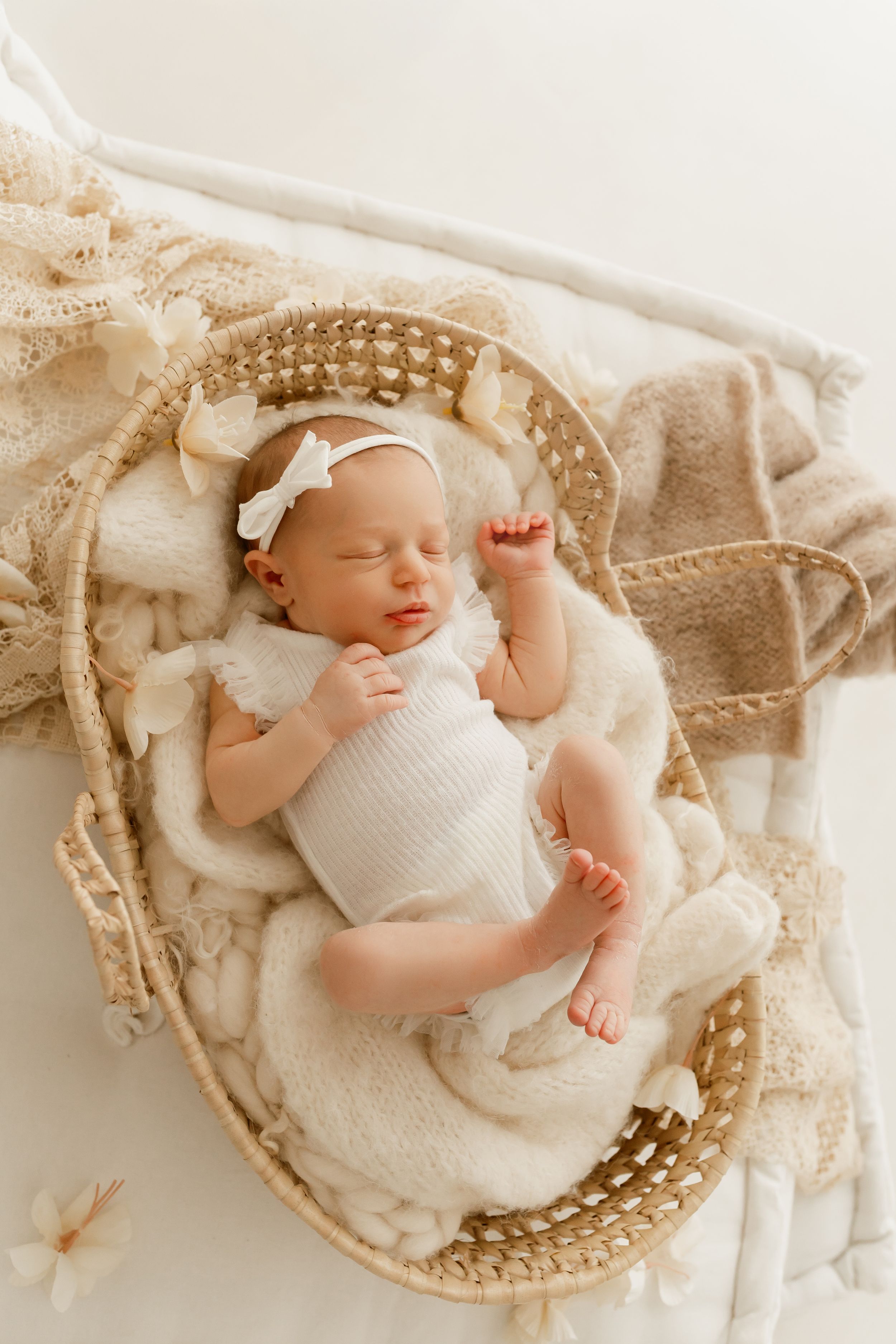 Newborn baby girl sleeping peacefully in a wicker Moses basket with cream blankets and a white bow headband.