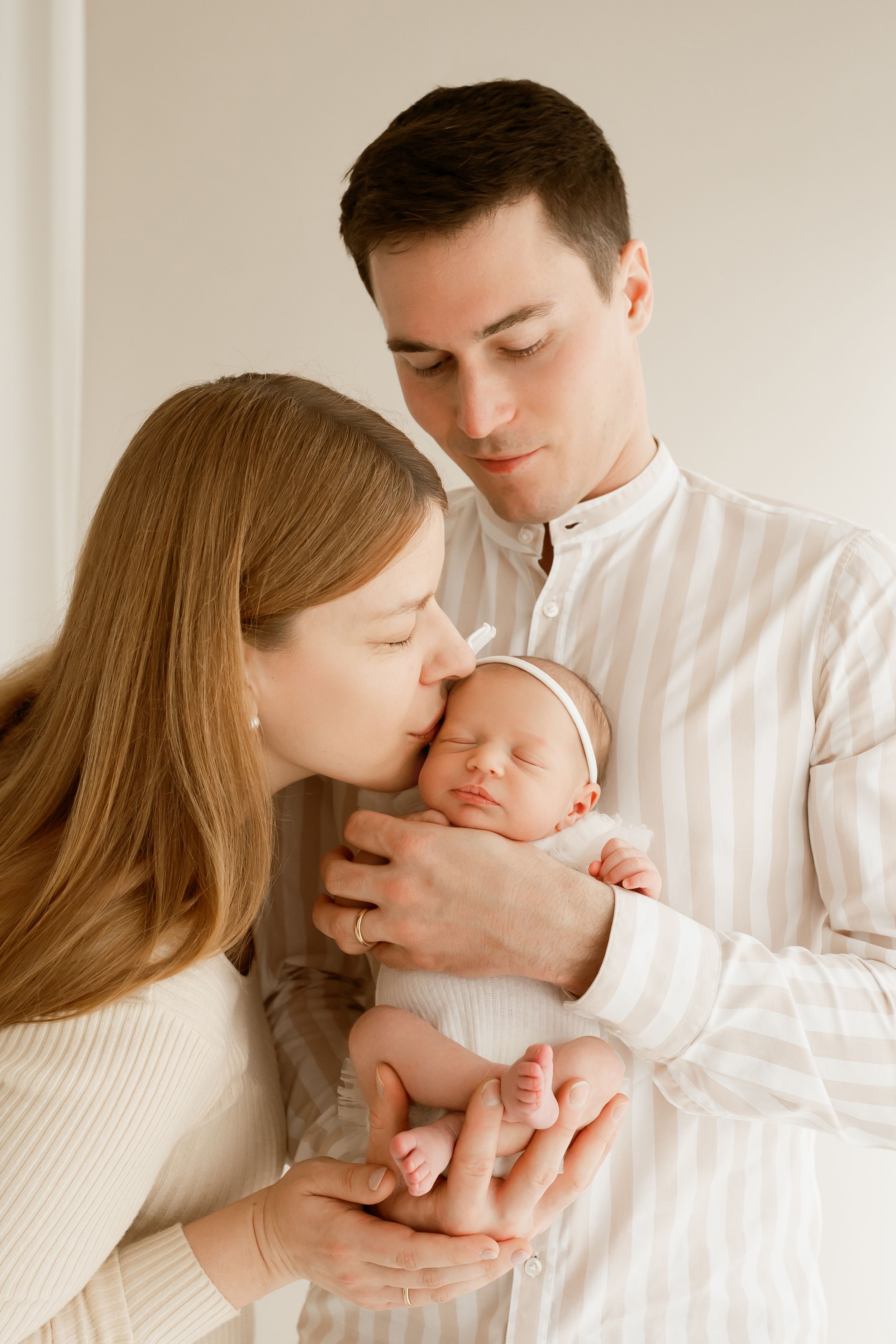 Young parents tenderly holding and kissing their sleeping newborn baby, dressed in white, in a bright room.