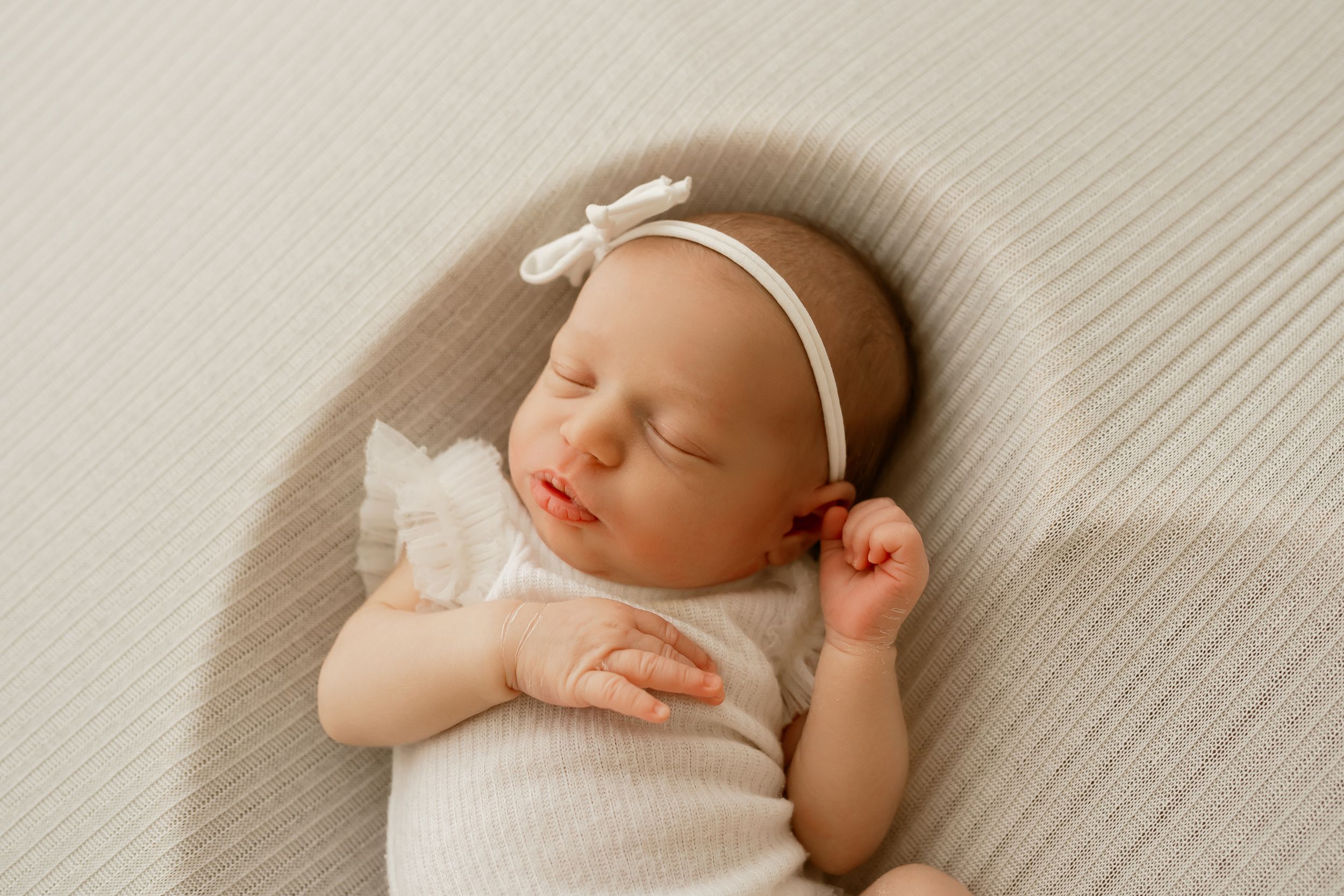 Peaceful newborn baby girl sleeping in white outfit and bow headband on soft neutral background.