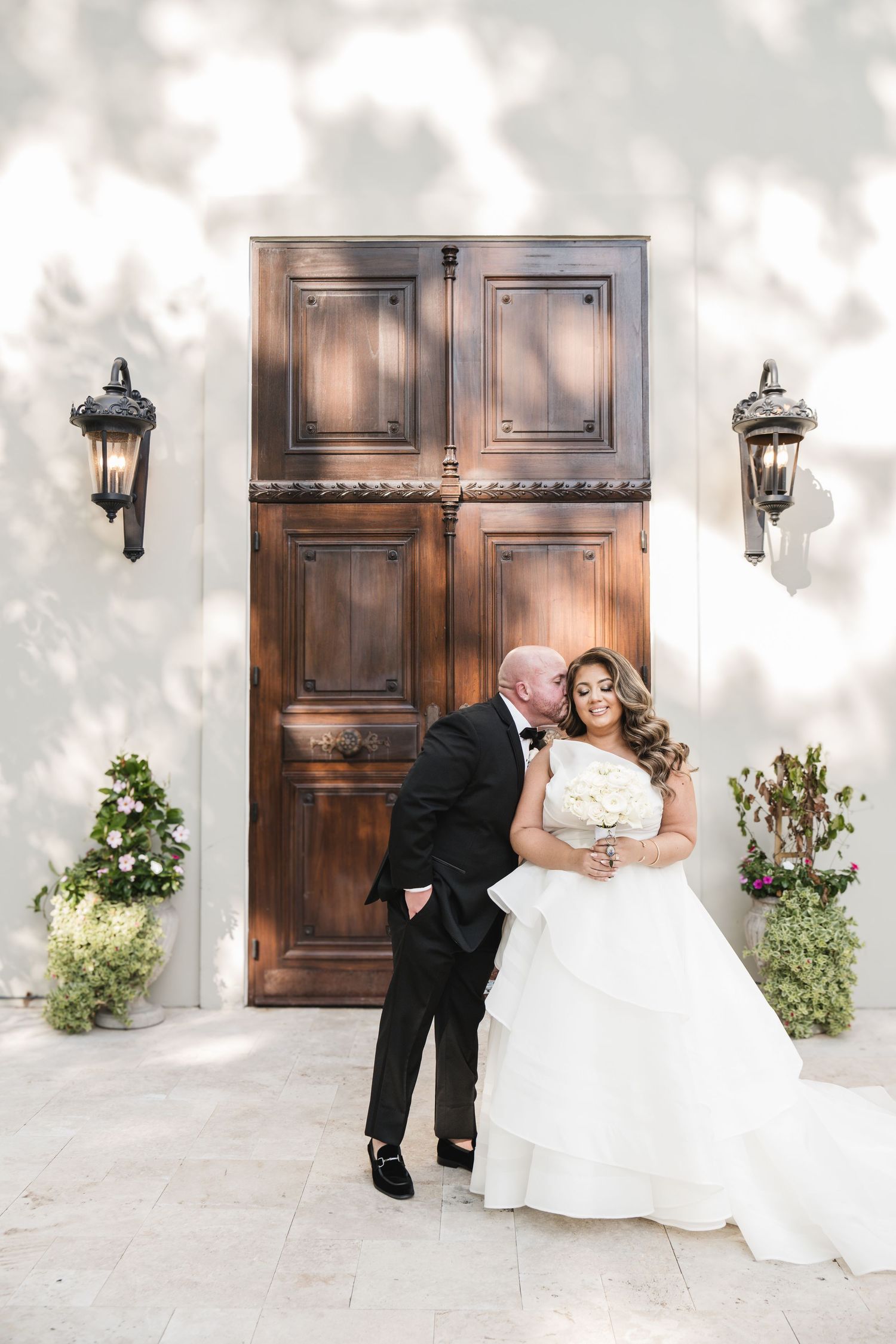 Bride and groom pose romantically before ornate wooden doors, bride holding white bouquet in elegant tiered gown.