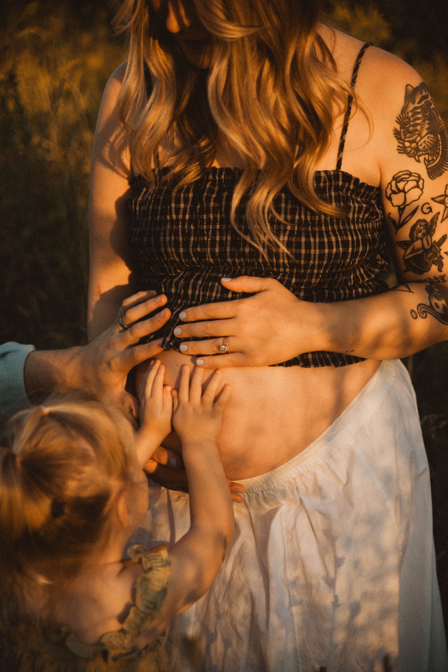 Pregnant woman with tattoos and blonde hair holds her baby bump while a young child touches her belly.