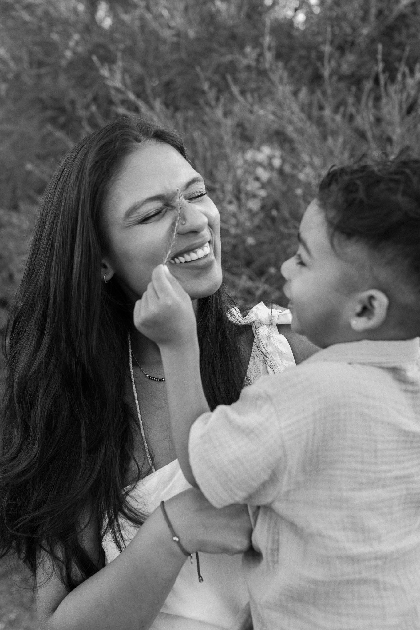 Black and white photo of a laughing woman and child playing together outdoors near lush greenery.