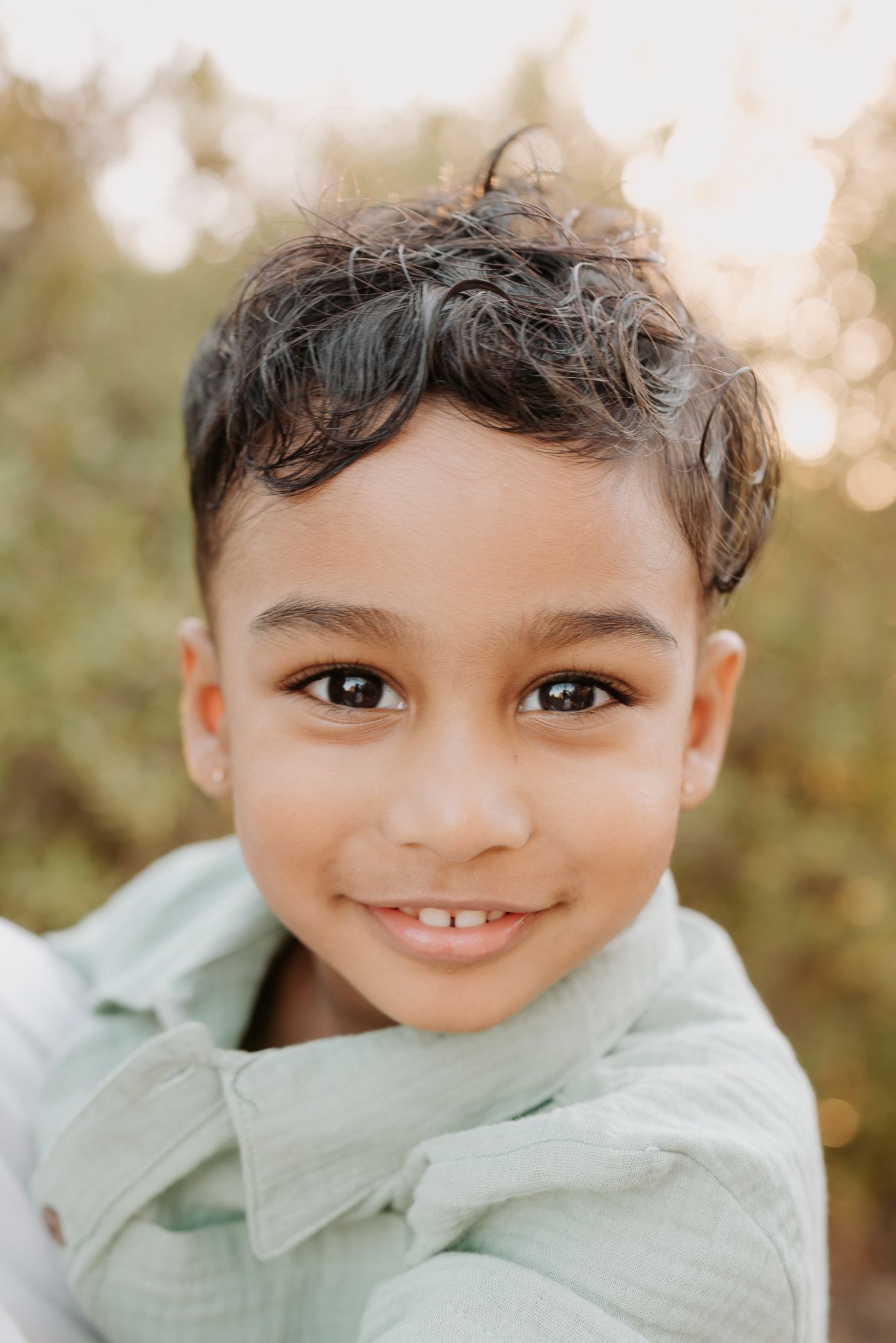 Smiling young boy with curly dark hair wearing a light green shirt outdoors with bokeh background.