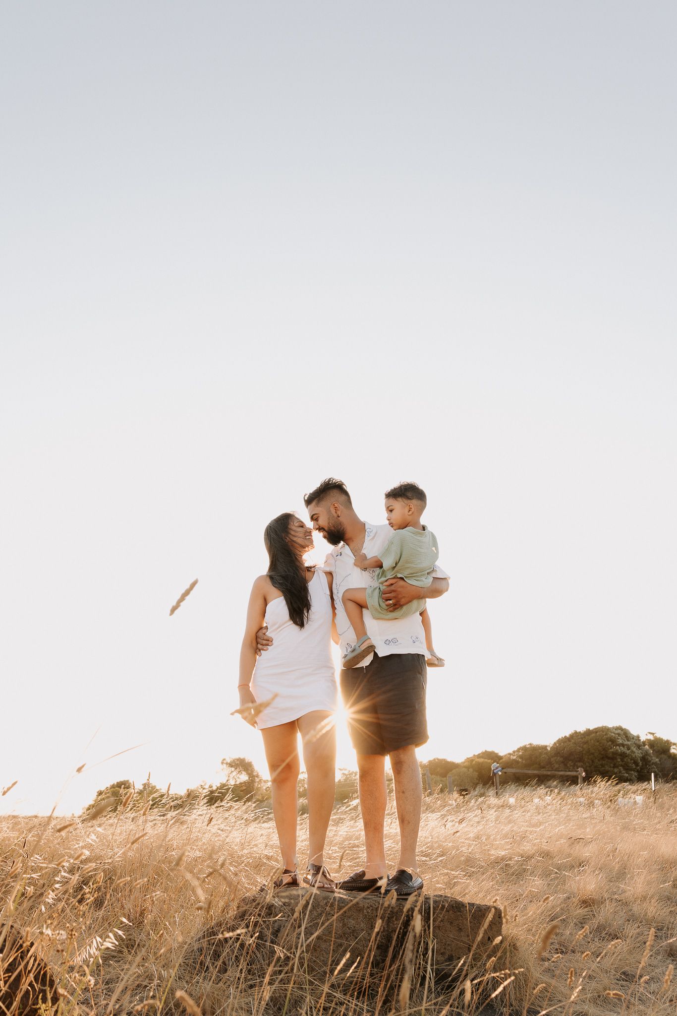 A couple sharing a kiss in a golden field at sunset, with the man holding their young child in his arms.