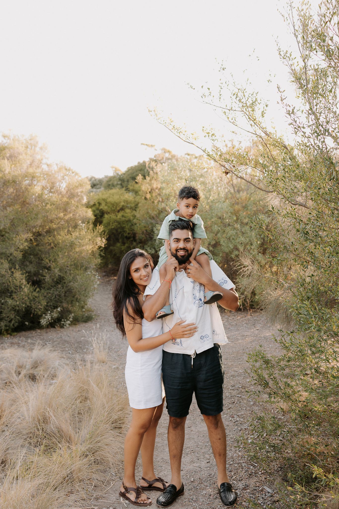 Family of three posing outdoors on a dirt path, mom in white dress, dad carrying young child on shoulders, surrounded by greenery.