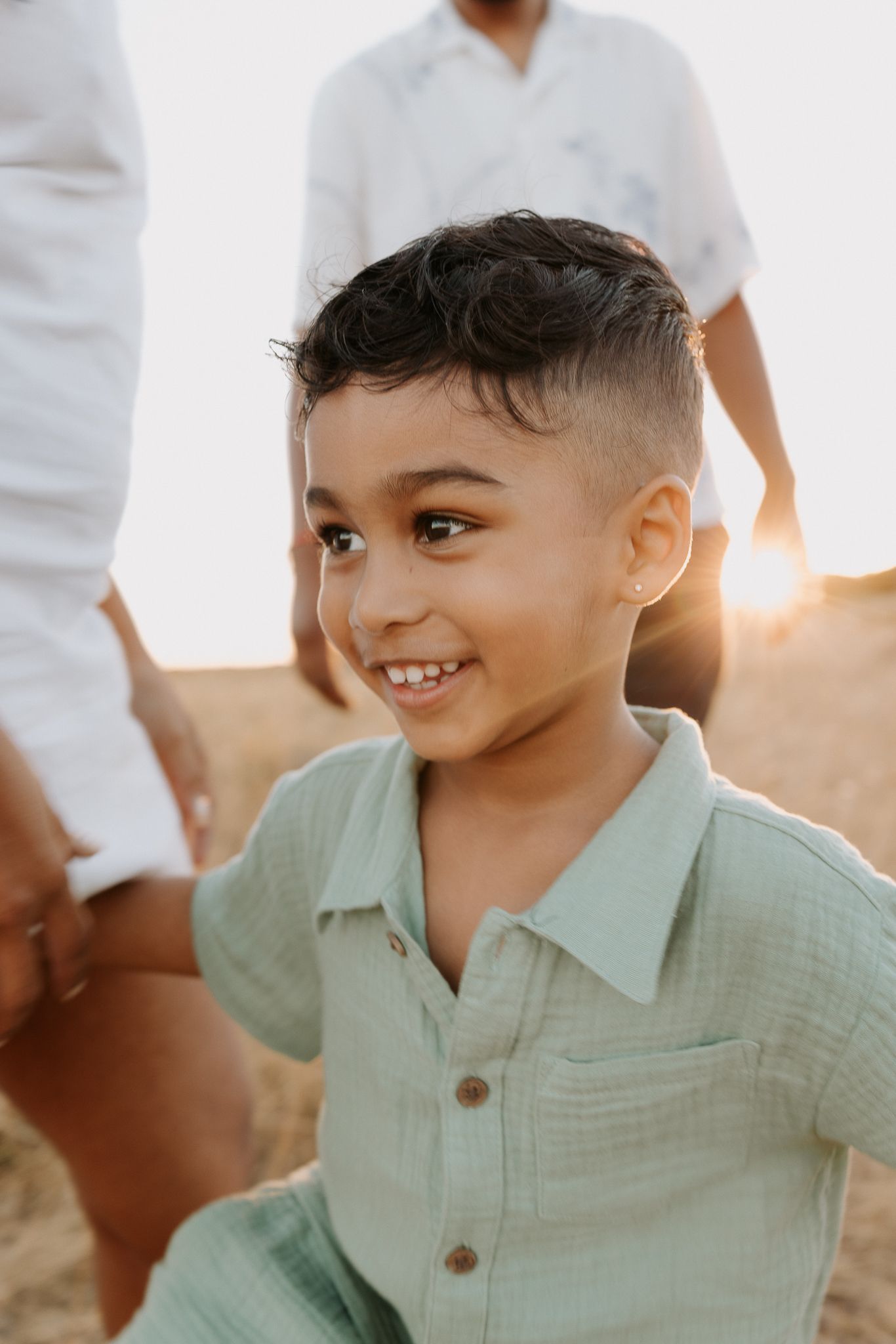 A smiling young boy in a sage green button-up shirt enjoys a sunny outdoor moment with family in the golden light.