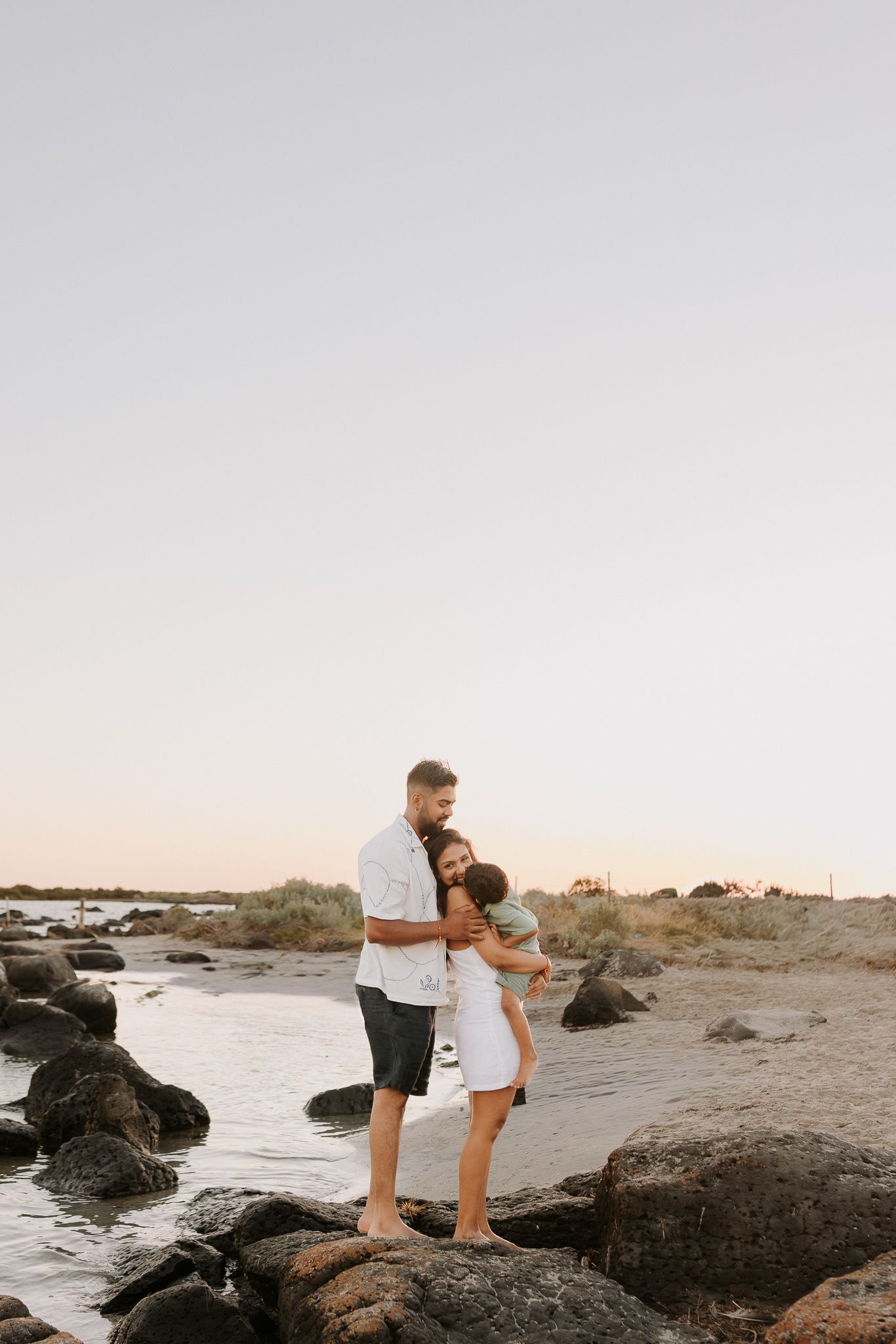 A couple embracing on rocky beach at sunset, woman in white dress holding baby, man in white shirt and dark shorts.