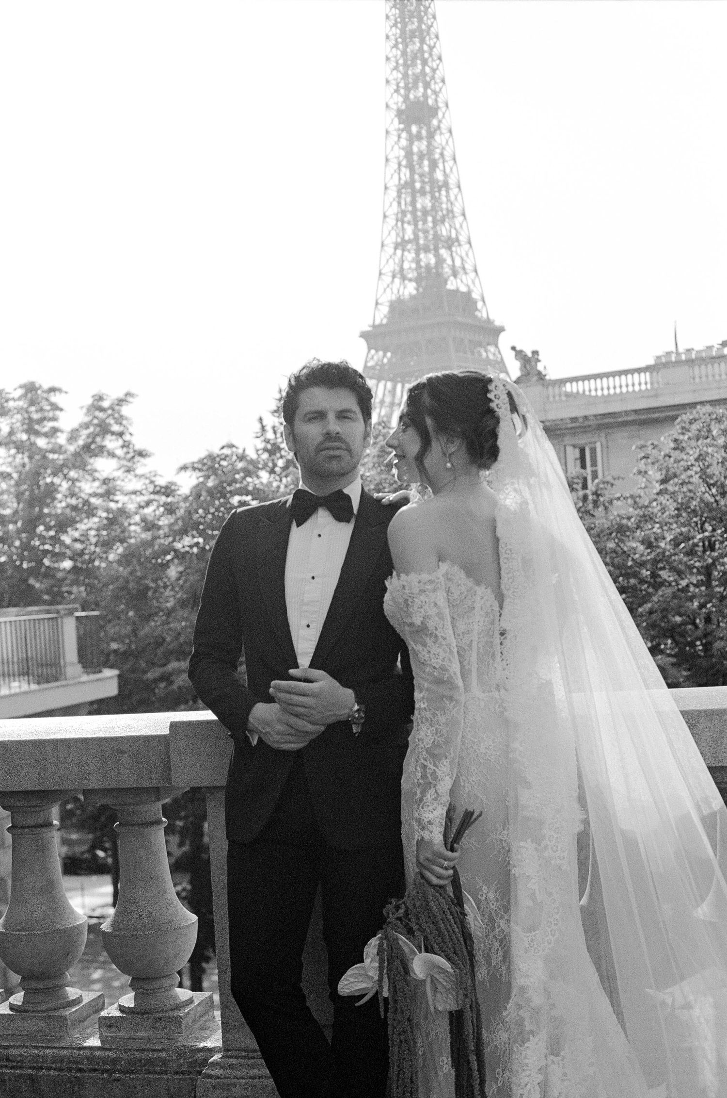 Black and white wedding photo of couple in formal attire posing on balcony with Eiffel Tower in Paris background.