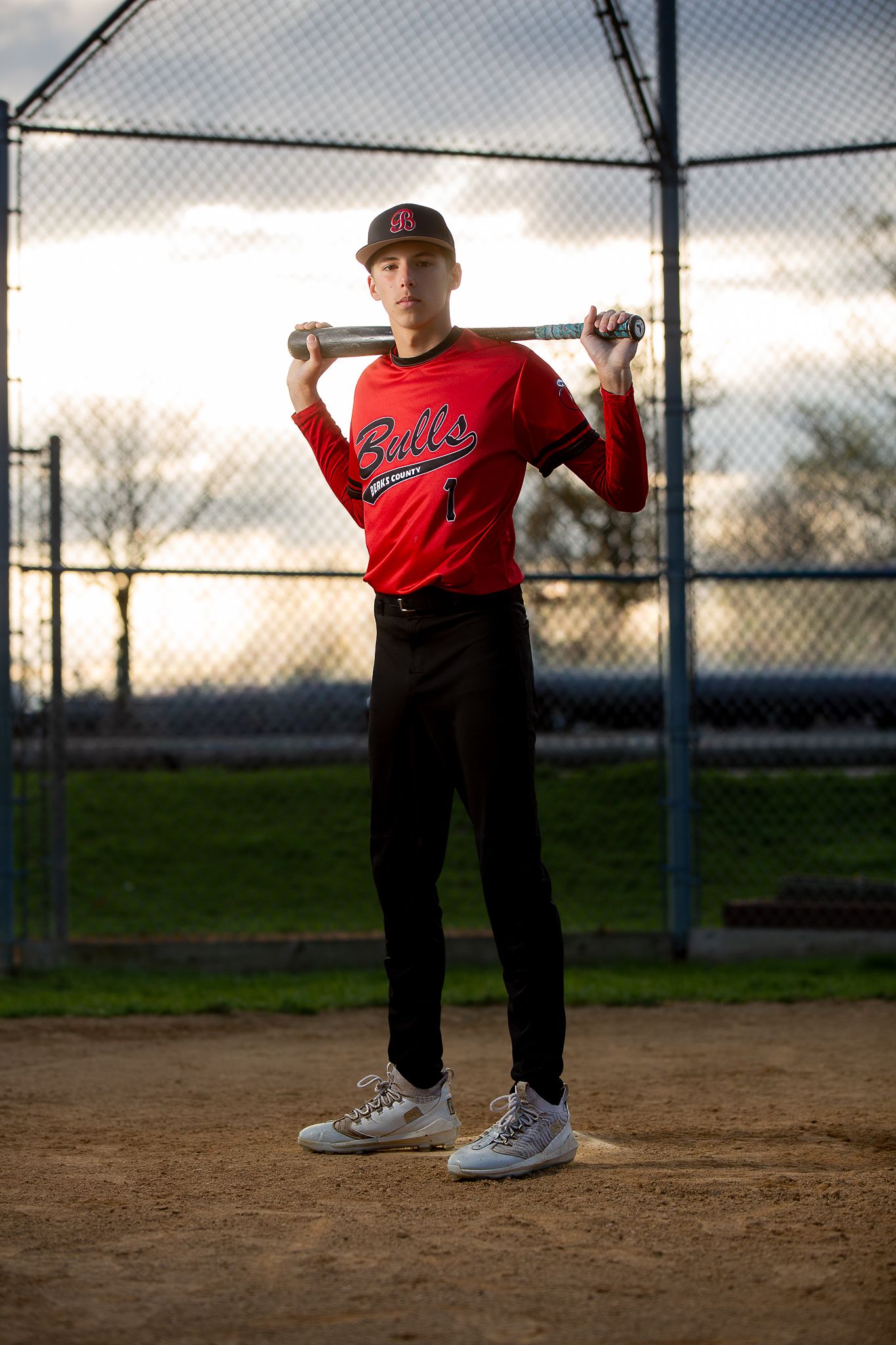 Young baseball player in red Bulls jersey poses with bat on shoulders at a baseball diamond during golden hour.